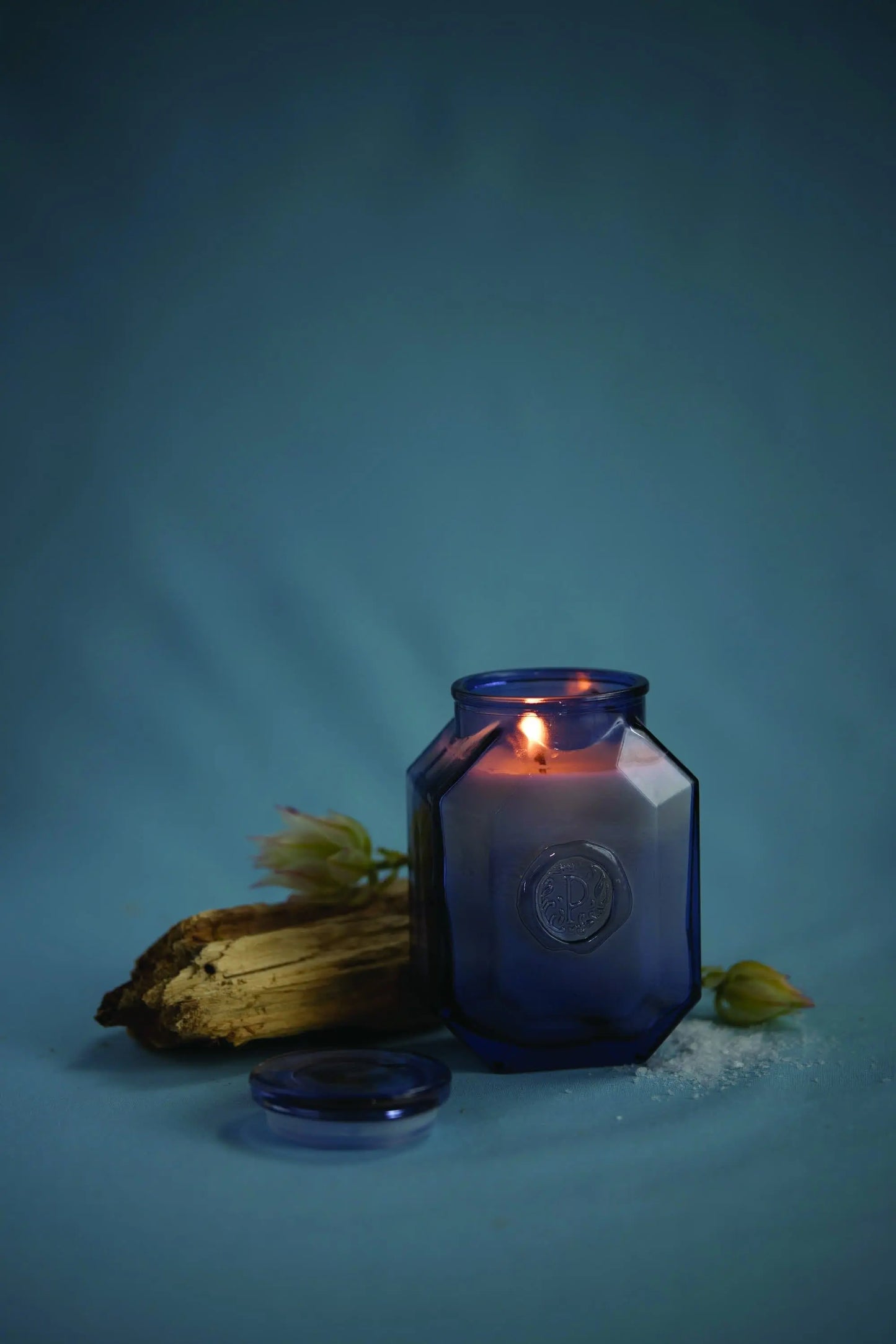 The Botanica Candle - Sea Salt + Amber from the Luxurious Botanica Collection glows in a blue glass jar beside wood and a flower bud, highlighting the collection’s elegance and refined glass vessels.