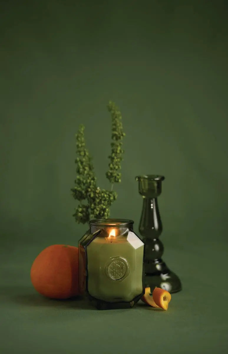 A lit Botanica Candle - Wild Neroli in a glass jar sits beside a green glass candlestick, a peach, lush green foliage, and peach slices, arranged on a vibrant green background.