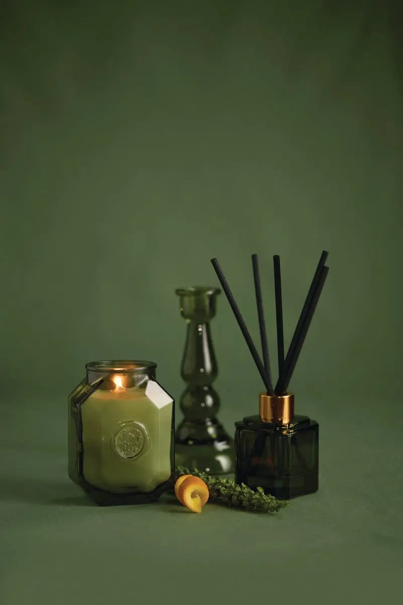 A lit green candle, a green glass candlestick holder, the Botanica Glass Diffuser - Wild Neroli in black, and small decorative items are displayed on a green surface with a matching background.