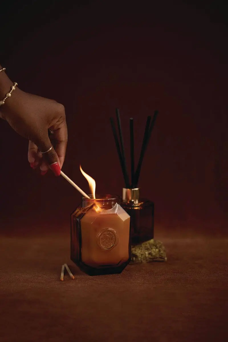 A hand lights a candle in a dark glass jar with a match. Behind it, the Botanica Glass Diffuser - Leather + Smoke from the Luxurious Botanica Collection and dried flowers rest on a brown surface, evoking rich leather and smoky notes.