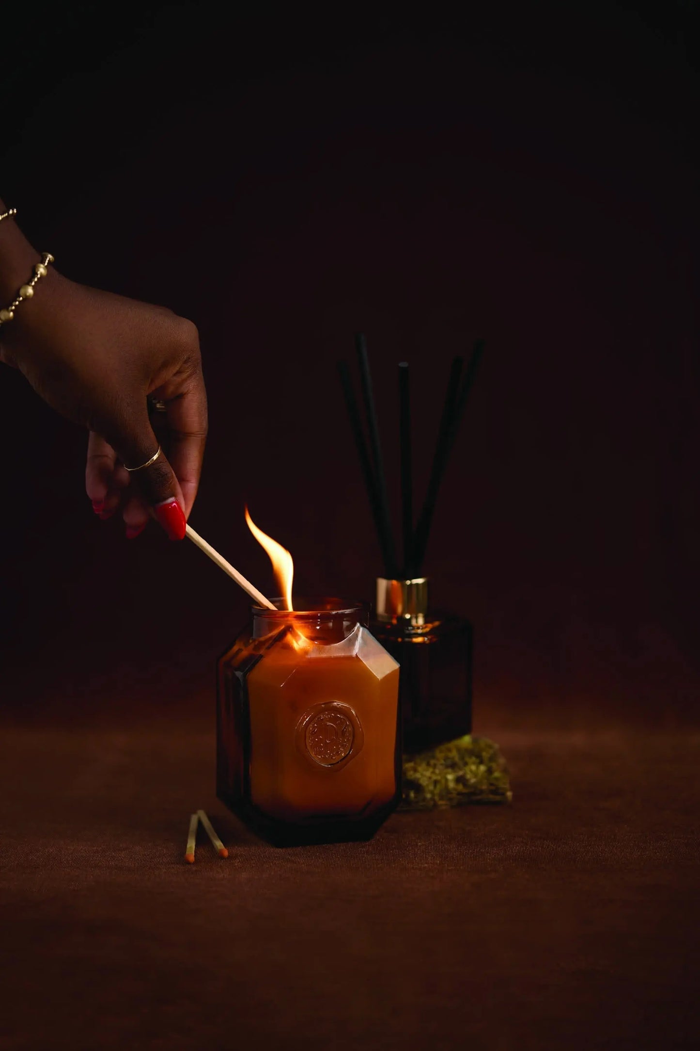 A hand lights the Botanica Candle - Leather + Smoke, housed in a brown glass jar, while black reed diffusers and dried herbs are visible in the background.