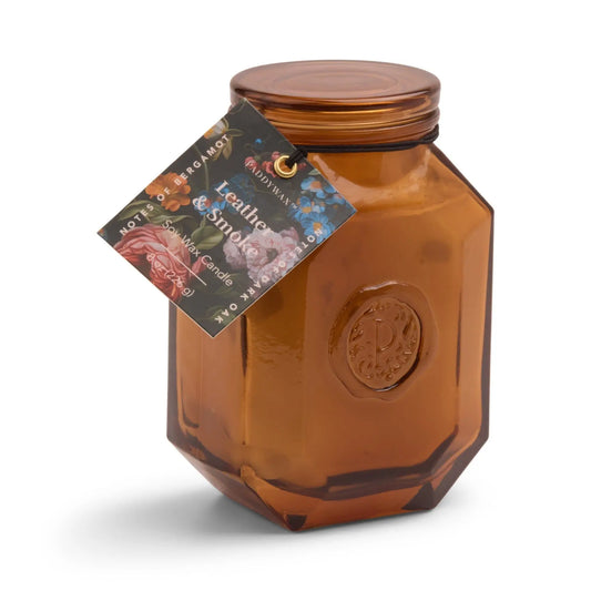 A brown, faceted glass jar from the Luxurious Botanica Collection with a screw-on lid and a floral tag labeled “Botanica Candle - Leather + Smoke.” Ideal for those who appreciate elegant candles and diffusers.
