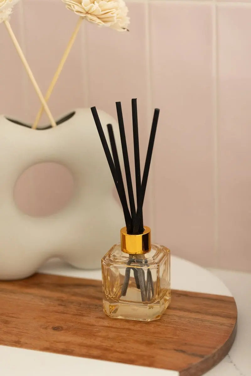 The Botanica Glass Diffuser - Cotton + Teak from the Luxurious Botanica Collection, featuring black reeds and a gold cap, sits on a wooden tray beside a white ceramic vase with dried flowers.