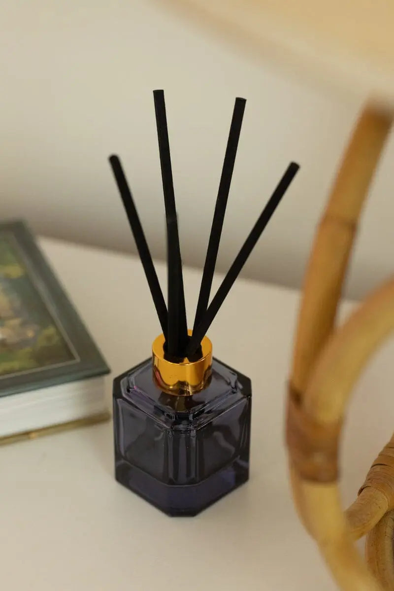 The Botanica Glass Diffuser - Sea Salt + Amber adds a refreshing scent to your space, featuring a black design with gold trim. It sits on a white surface beside a closed book and part of a wicker chair.
