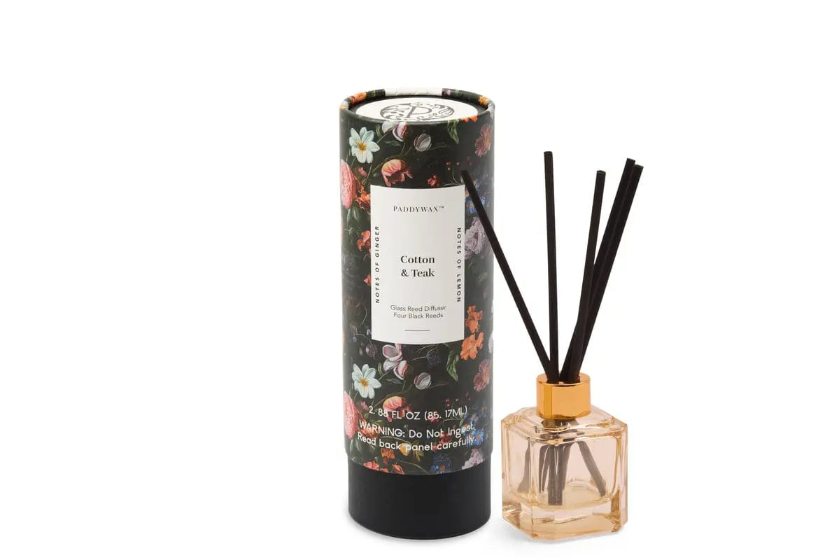 A floral-patterned tube labeled Botanica Glass Diffuser - Cotton + Teak stands beside a glass diffuser with black reeds and light liquid, part of the Luxurious Botanica Collection for an elegant, modern touch.