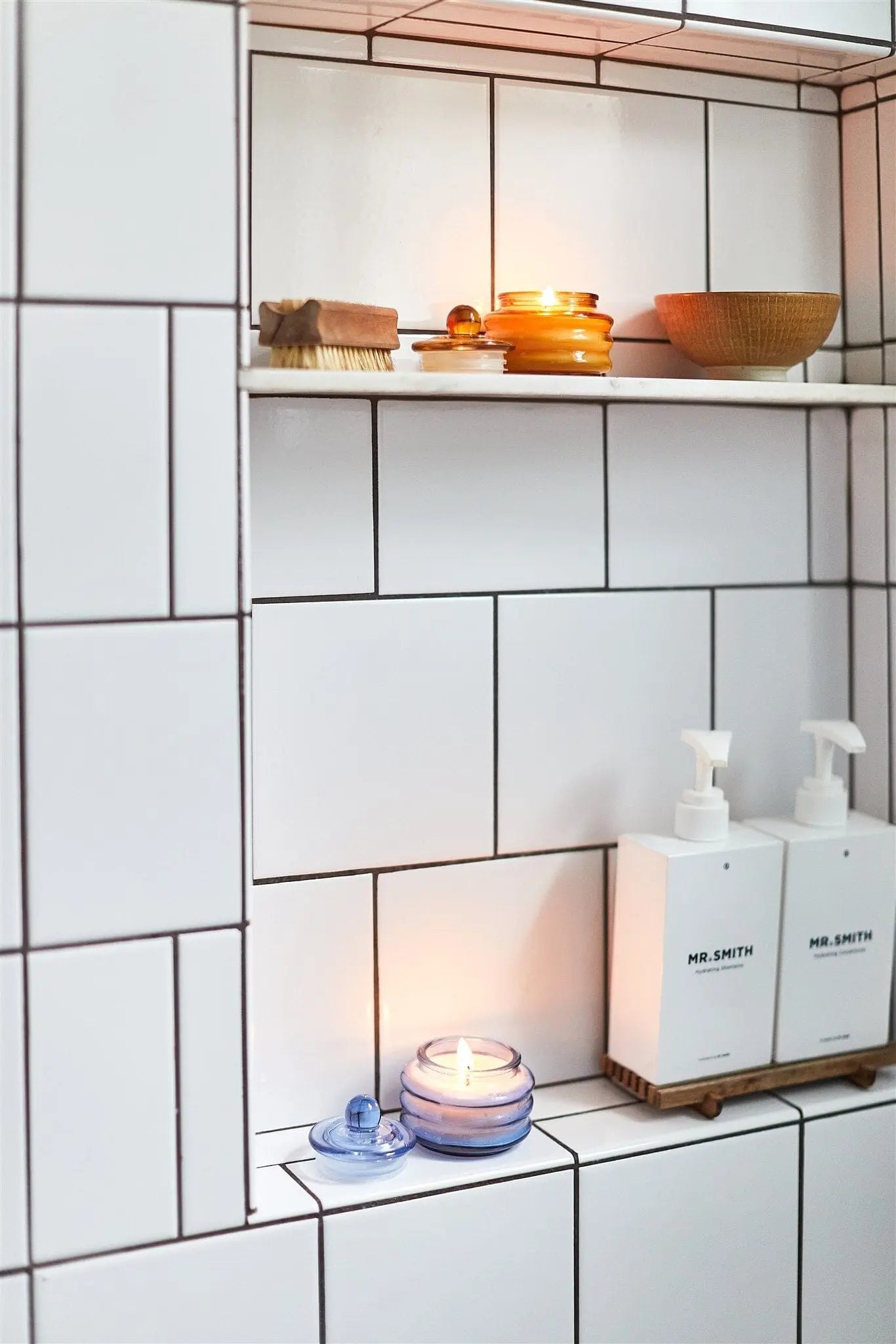 White tiled bathroom shelves with a Beam 3oz Glass Candle - Pepper + Plum casting ambient light, two MR.SMITH pump bottles, a small wooden bowl, brush, and stacked glass jars—ideal for indulging in luxurious scents.