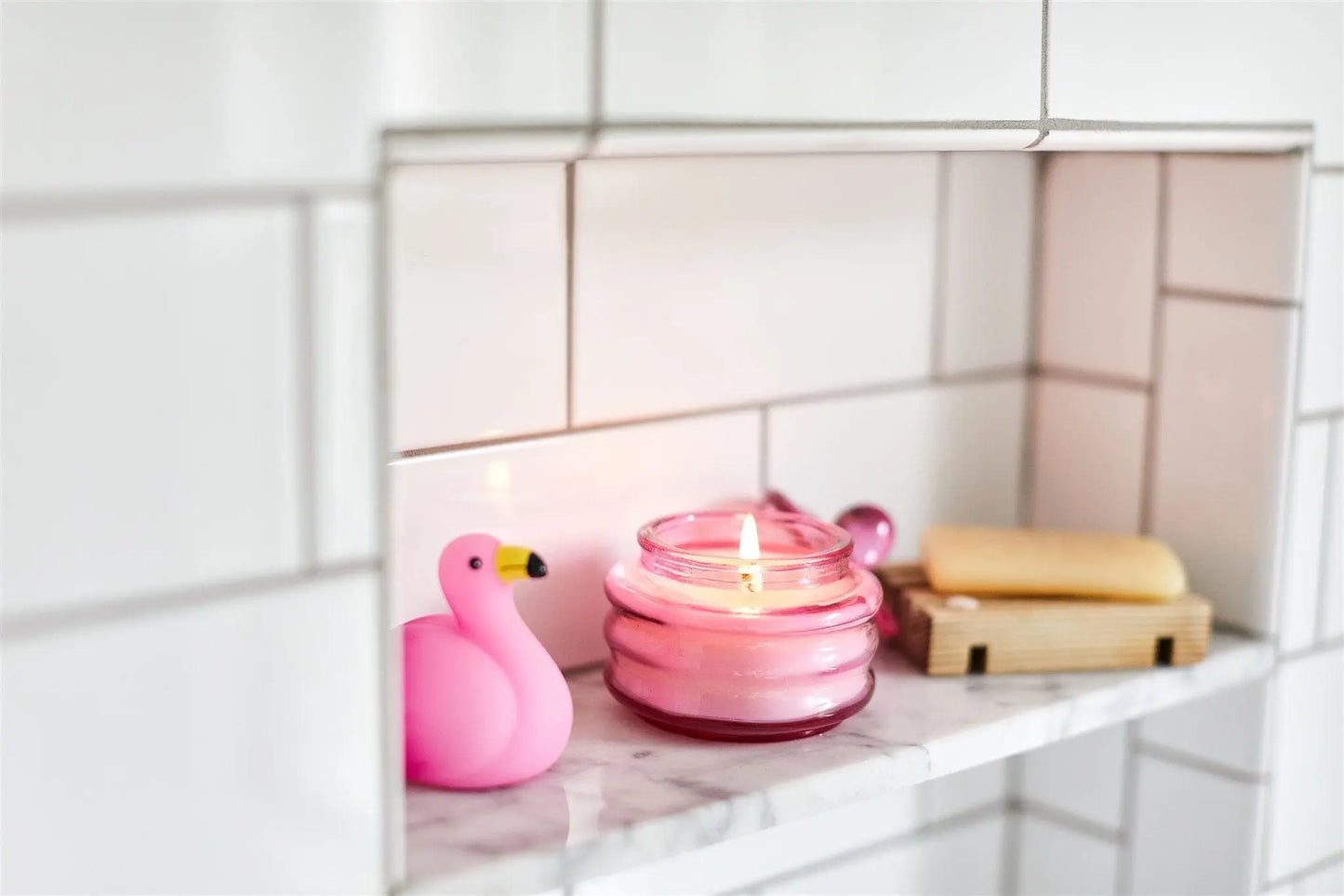 A Beam 3oz Glass Candle in Desert Peach emits a soft fragrance as it sits with a pink rubber flamingo, a bar of soap on a wooden dish, and a spoon rest atop a marble shelf against white tiled walls.
