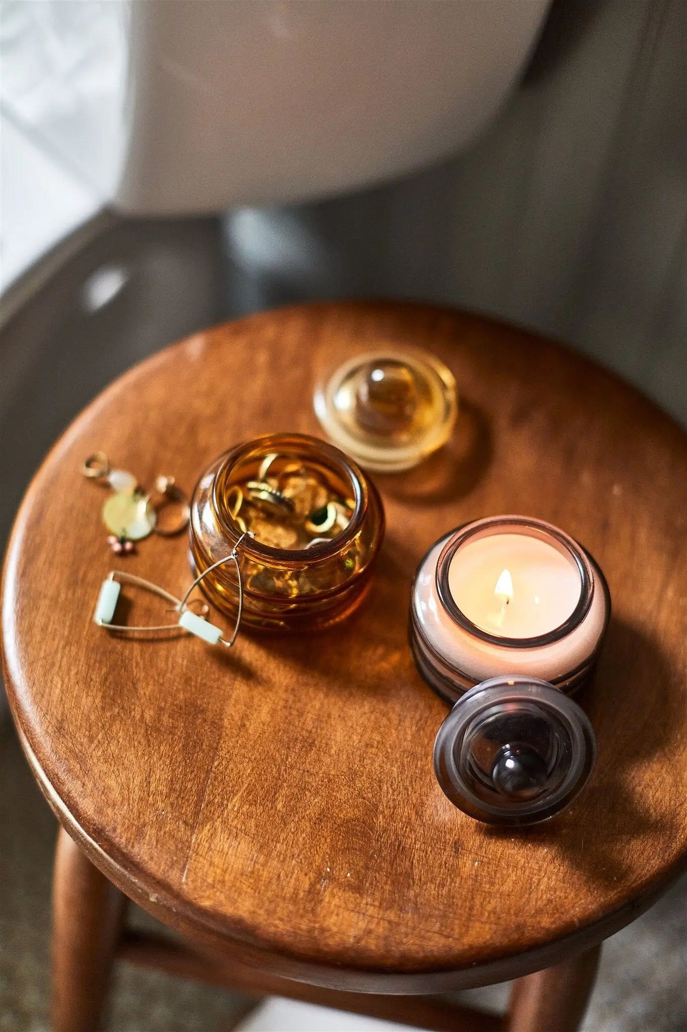 A wooden stool holds a Beam 3oz Glass Candle - Baltic Ember casting ambient light, an open jar of capsules, a small lidded jar, and a pair of gold earrings.