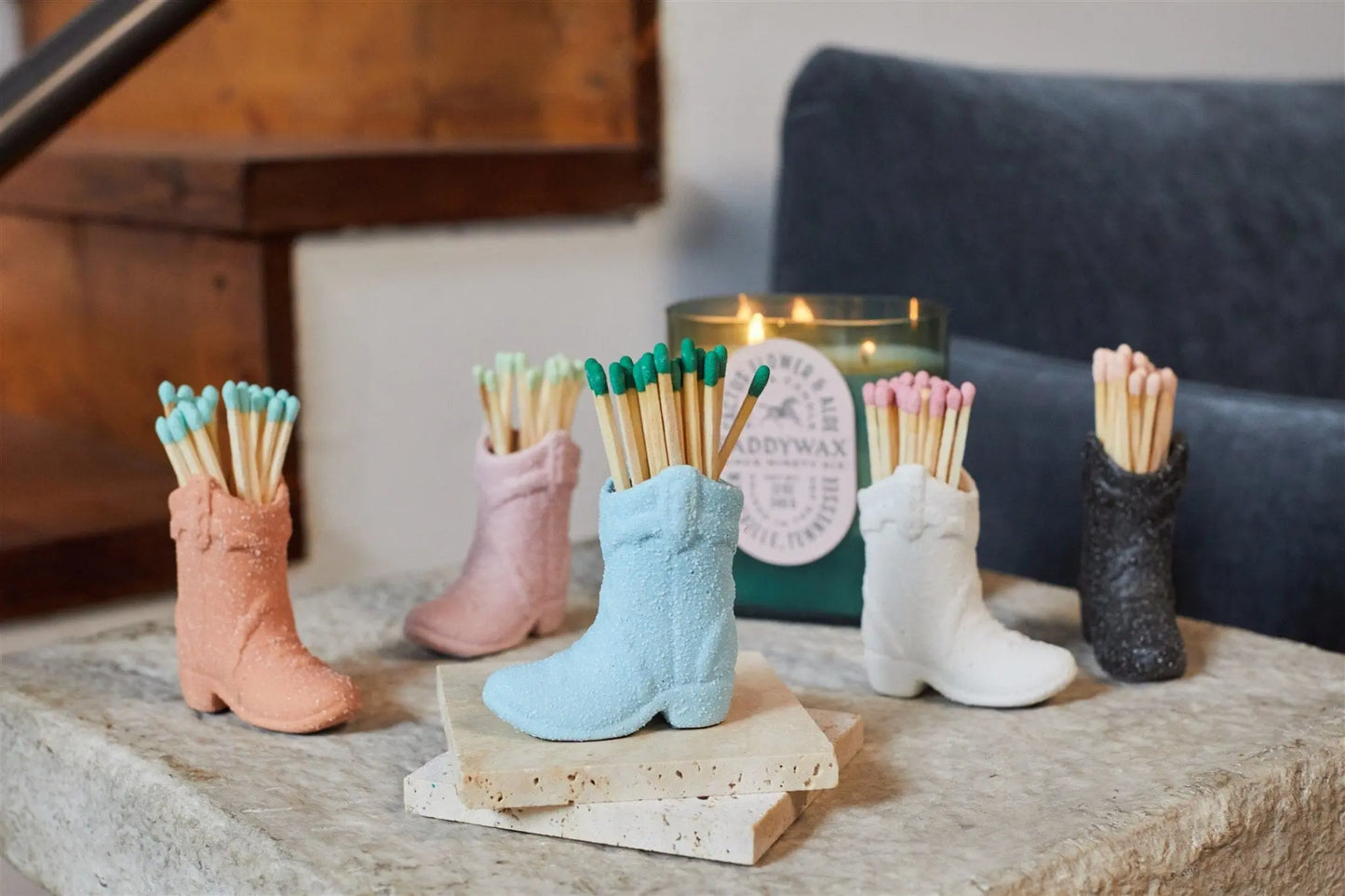 The Cowboy Boot Match Holder - White adds a Western vibe as it holds matches on a stone surface next to a lit candle.