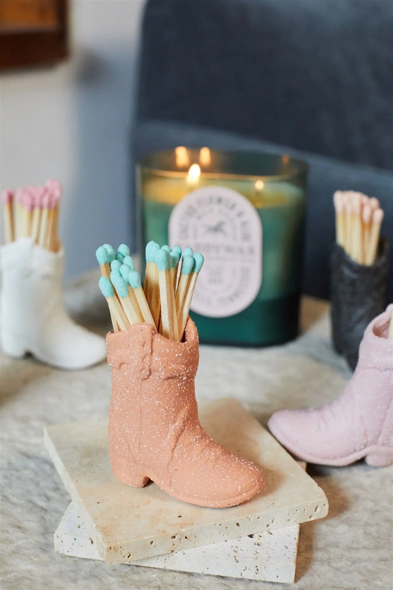 The Cowboy Boot Match Holder - Terracotta sits on a stone surface, filled with colorful matches, adding a Western vibe as a candle glows in the background.