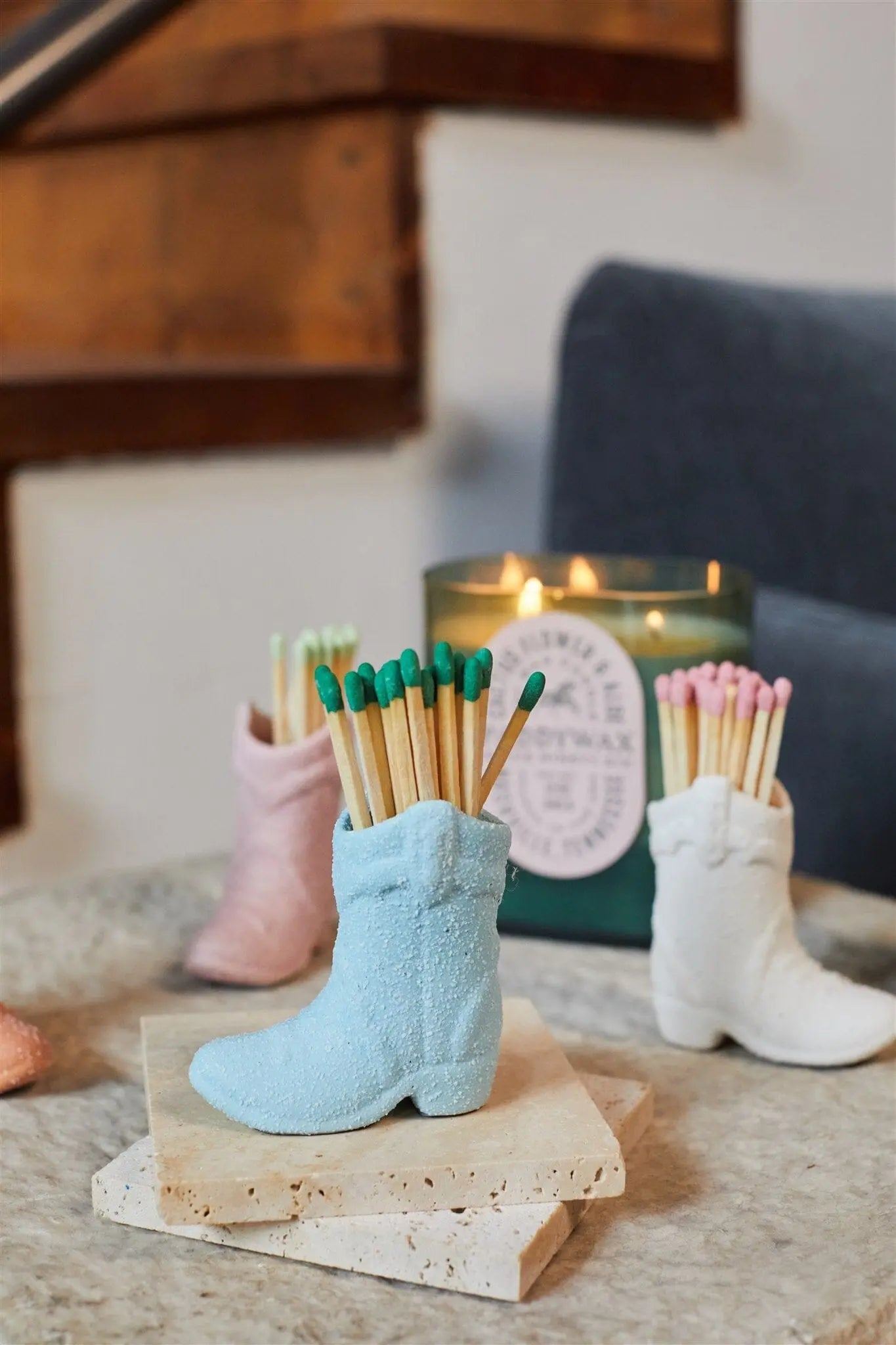 A Cowboy Boot Match Holder in light blue, filled with colored-tipped matches, rests on a stone surface near a lit candle and staircase—perfect as a ceramic boot accessory for vintage-inspired home decor.
