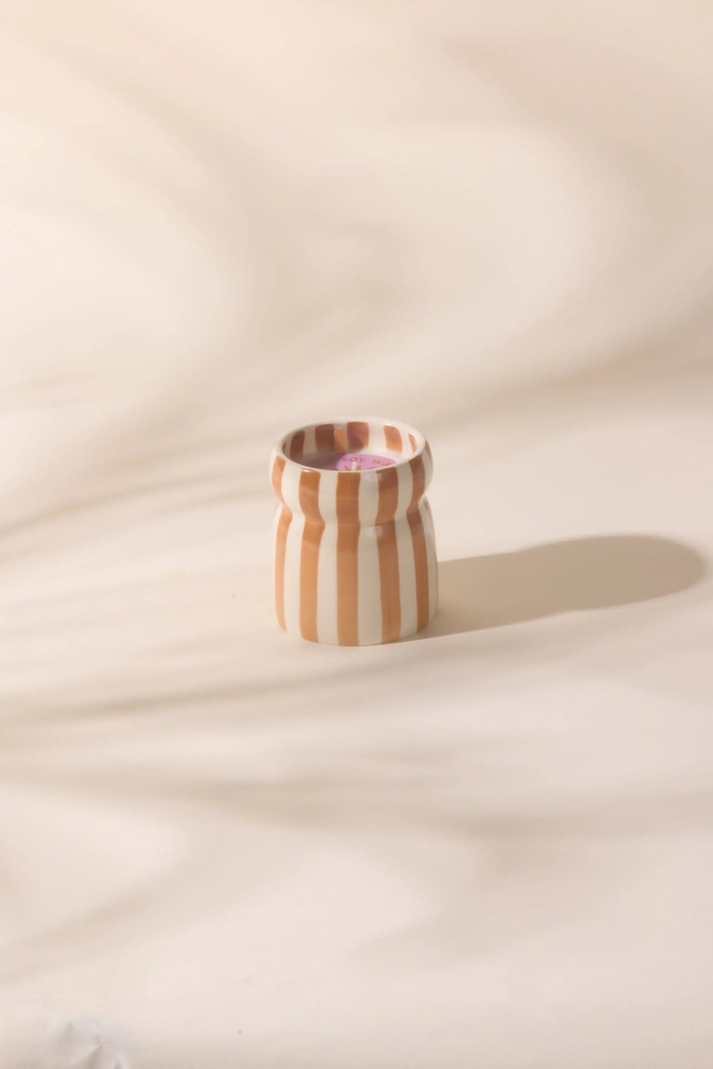 The Cabana Rust Candle - Sienna Sunset, a small food-safe ceramic piece with vertical beige and white stripes, is shown on a light surface with soft shadows in the background.