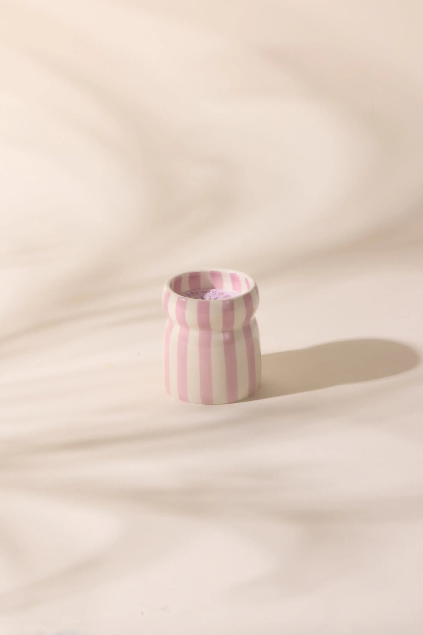 The Cabana Lavender Candle - Grecian Sand features a small ceramic vessel with vertical pink and white stripes, casting a soft shadow—perfect for brightening your space or bringing coastal charm to any room.
