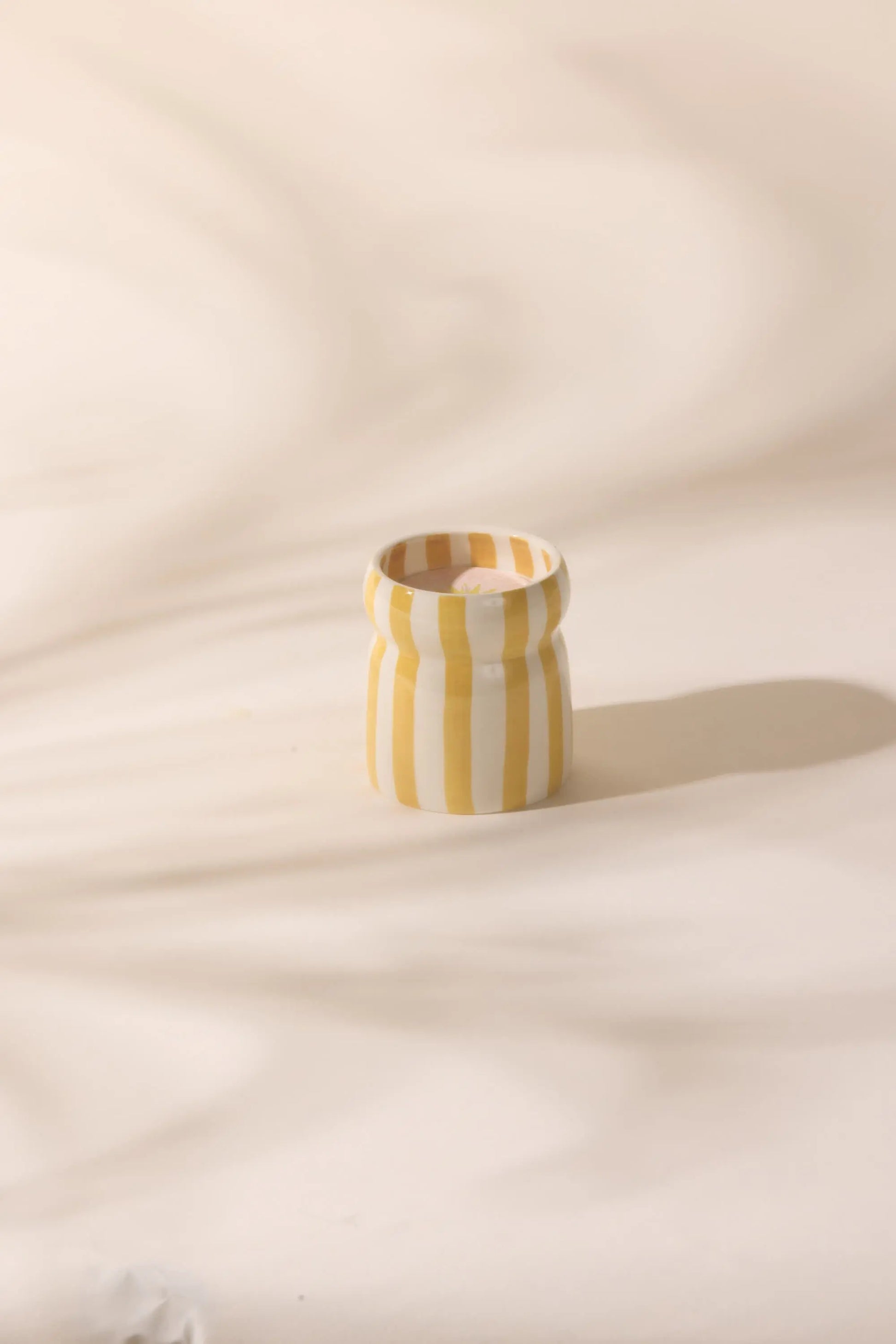 The Cabana Ceramic Candle - Gold Coast, featuring vertical yellow and white stripes, rests on a softly textured surface with a gentle shadow—capturing the relaxed charm of coastal getaways.