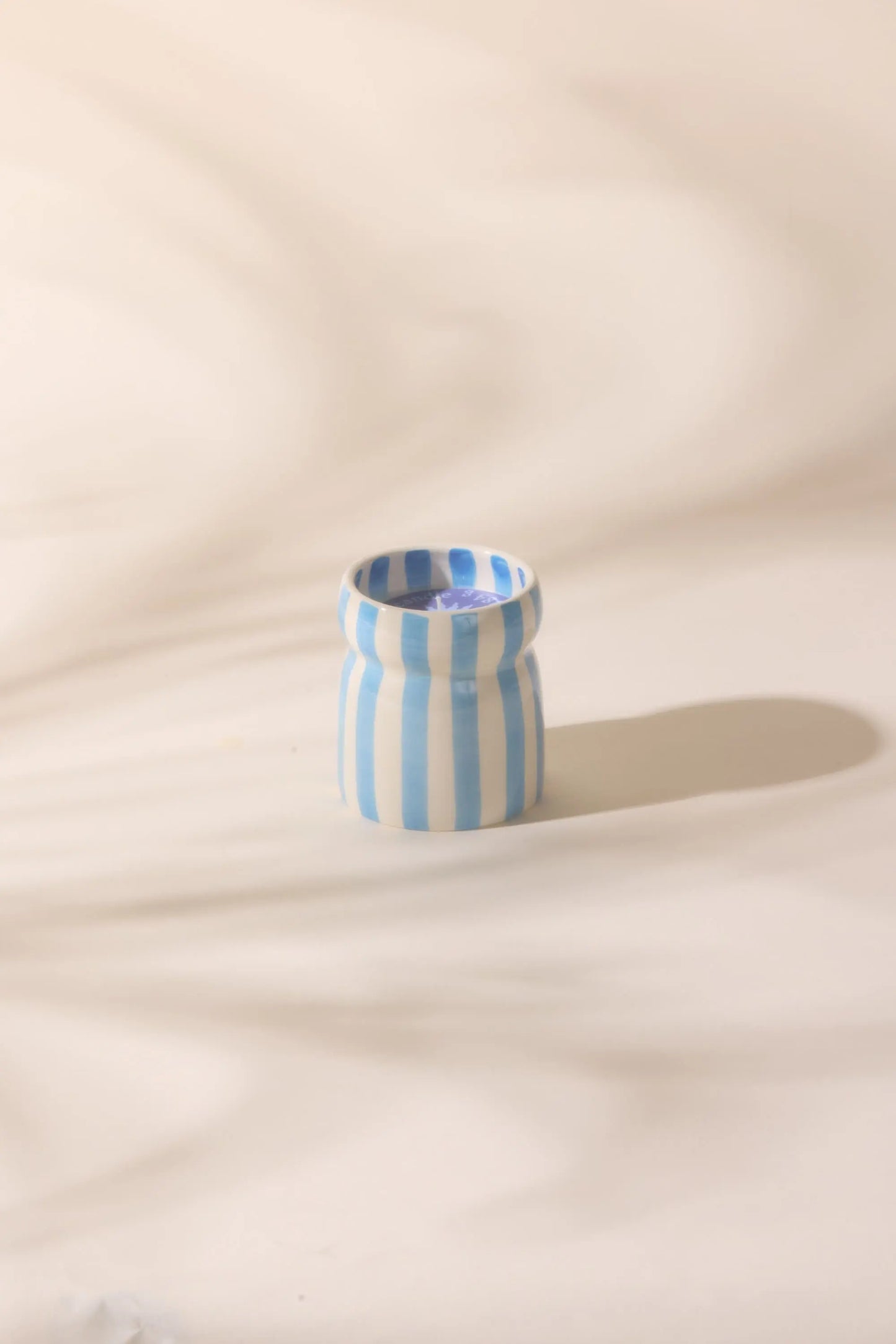The Cabana Blue Candle - Lost At Sea, featuring vertical blue and white stripes, rests on a smooth, light-colored surface and casts a gentle shadow evoking tranquil coastal vibes.