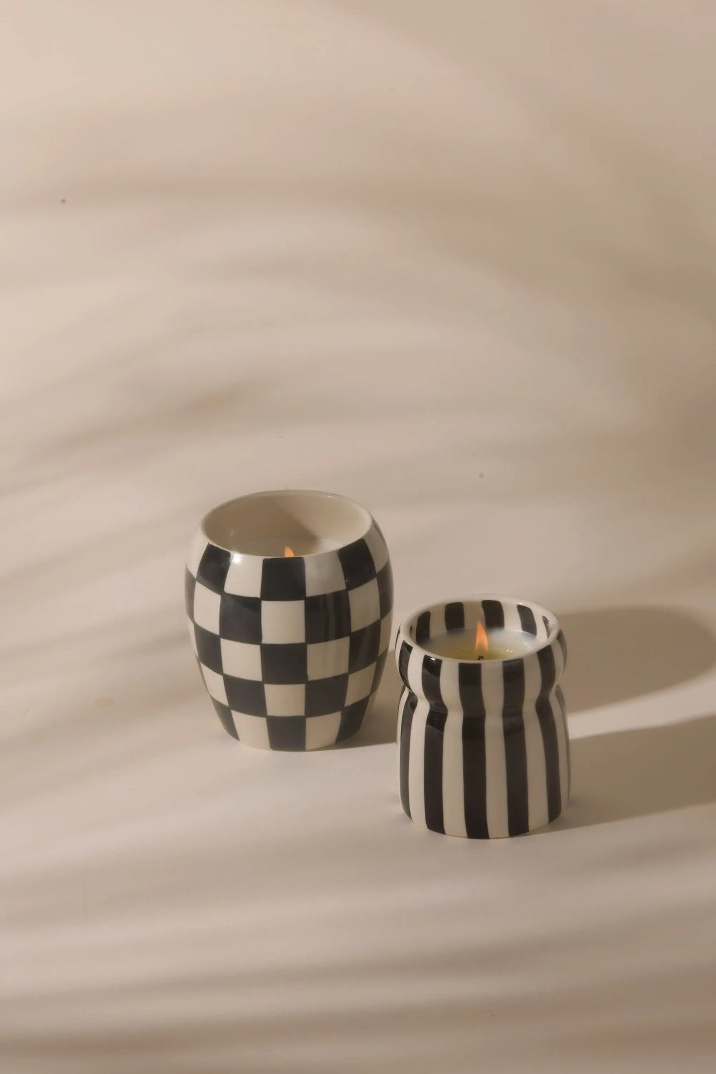 A lit, hand-painted Cabana Black Candle - Lava Stone sits in a black-and-white ceramic vessel on a smooth, light surface.