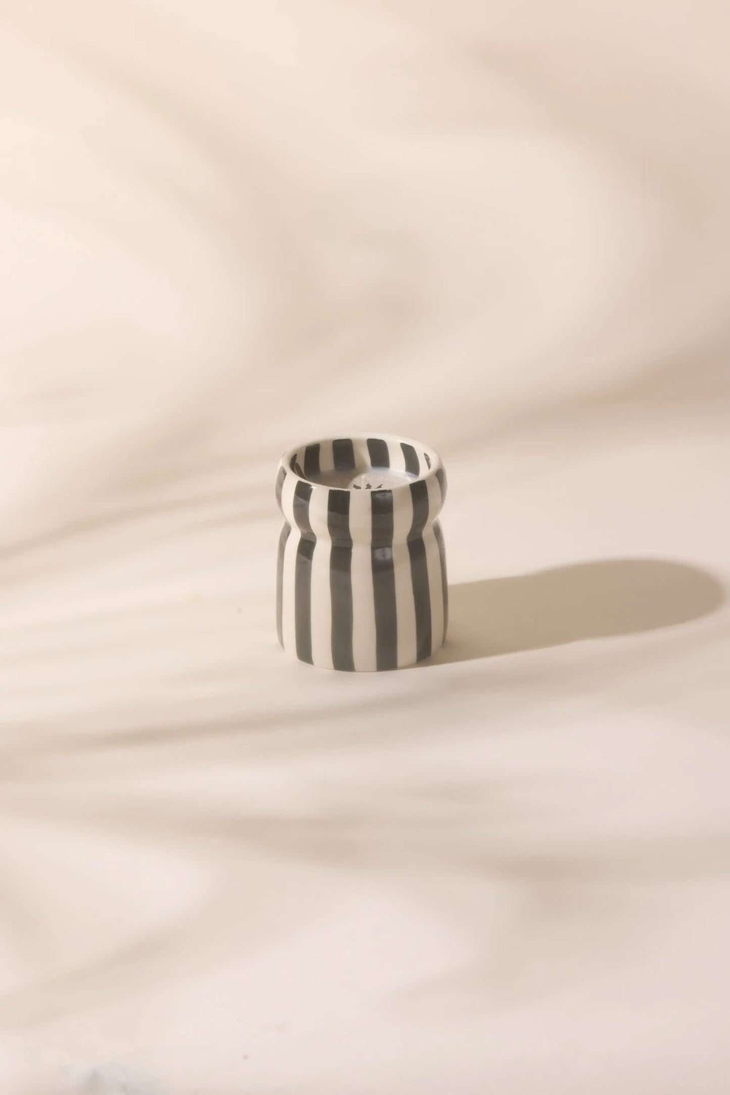 The Cabana Black Candle – Lava Stone, a small ceramic vessel with vertical black and white stripes, rests on a light, softly textured surface with its shadow extending to the right.