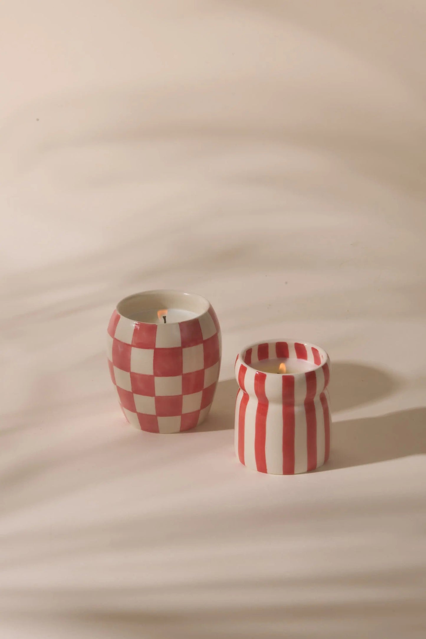 Two hand-painted Cabana Red Candle - Hibiscus Bay candles rest on a cream surface: one showcases red and white checks, the other vertical stripes. Perfect for adding a touch of Hibiscus Bay charm to any space.