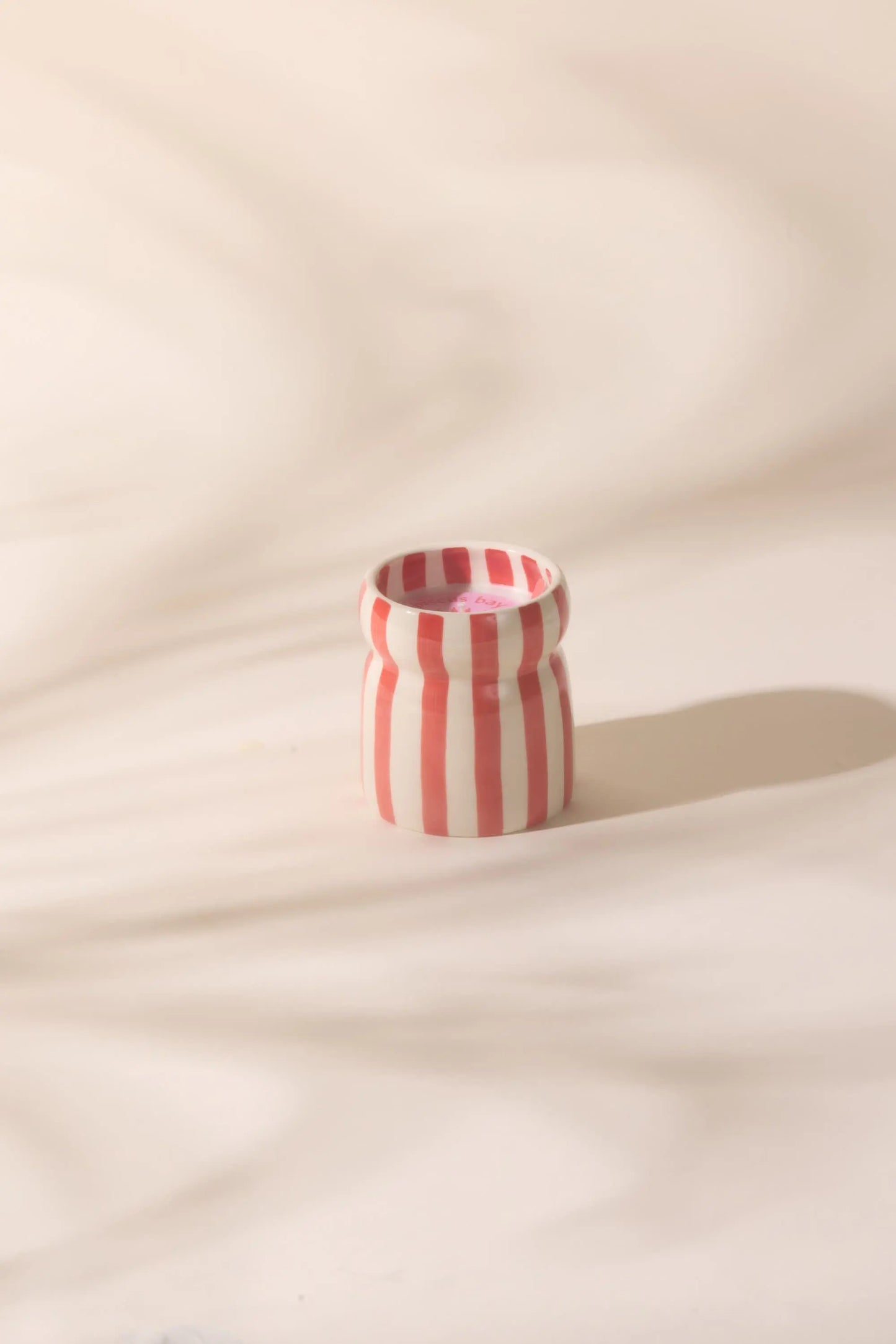The Cabana Red Candle - Hibiscus Bay, with vertical red and white stripes, sits on a light, softly textured surface and casts a shadow to the right.