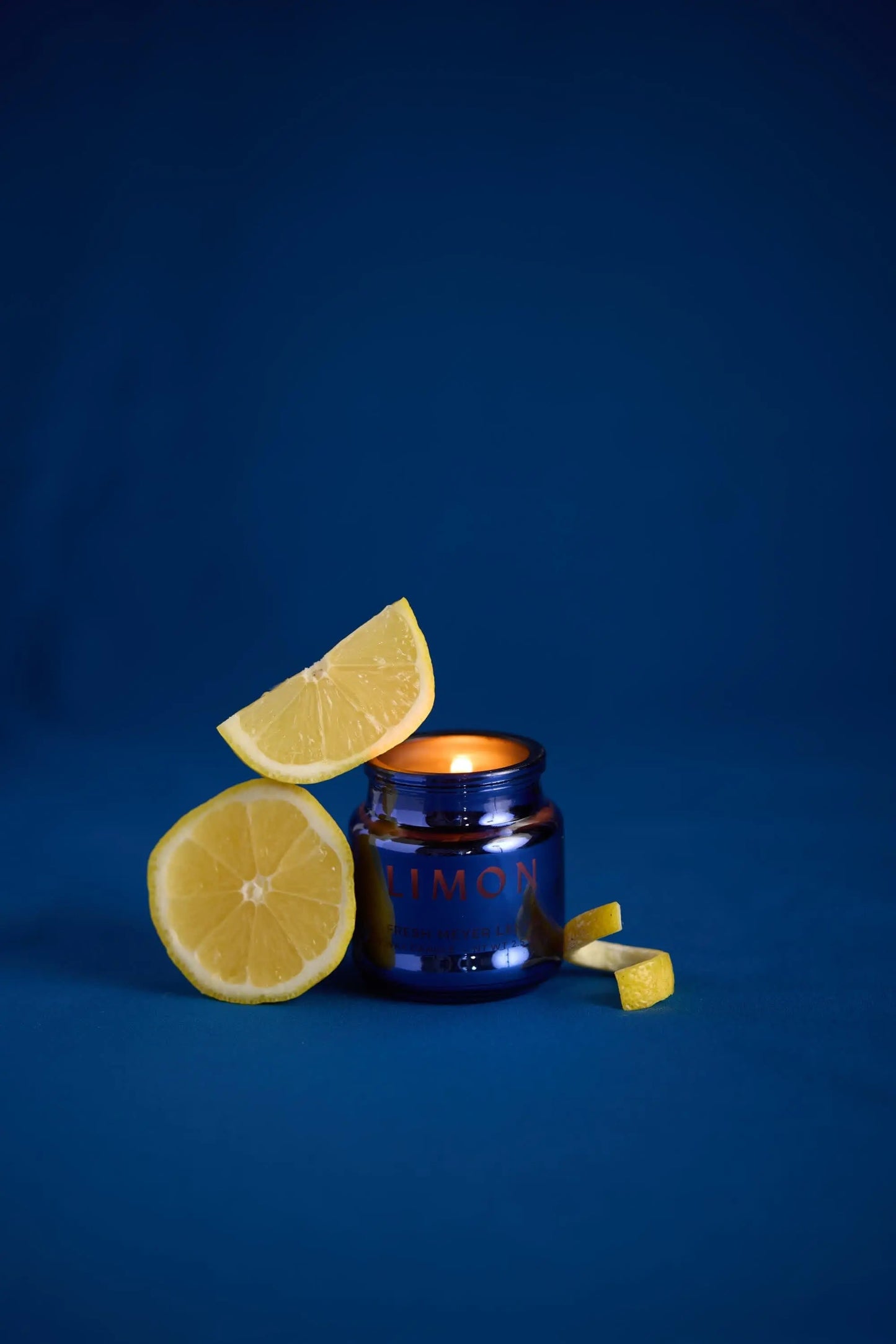The Chroma Candle - Meyer Lemon glows in a dark blue electroplated jar with LIMON on it, surrounded by lemon slices and peels on a solid blue background, offering a unique scent experience.