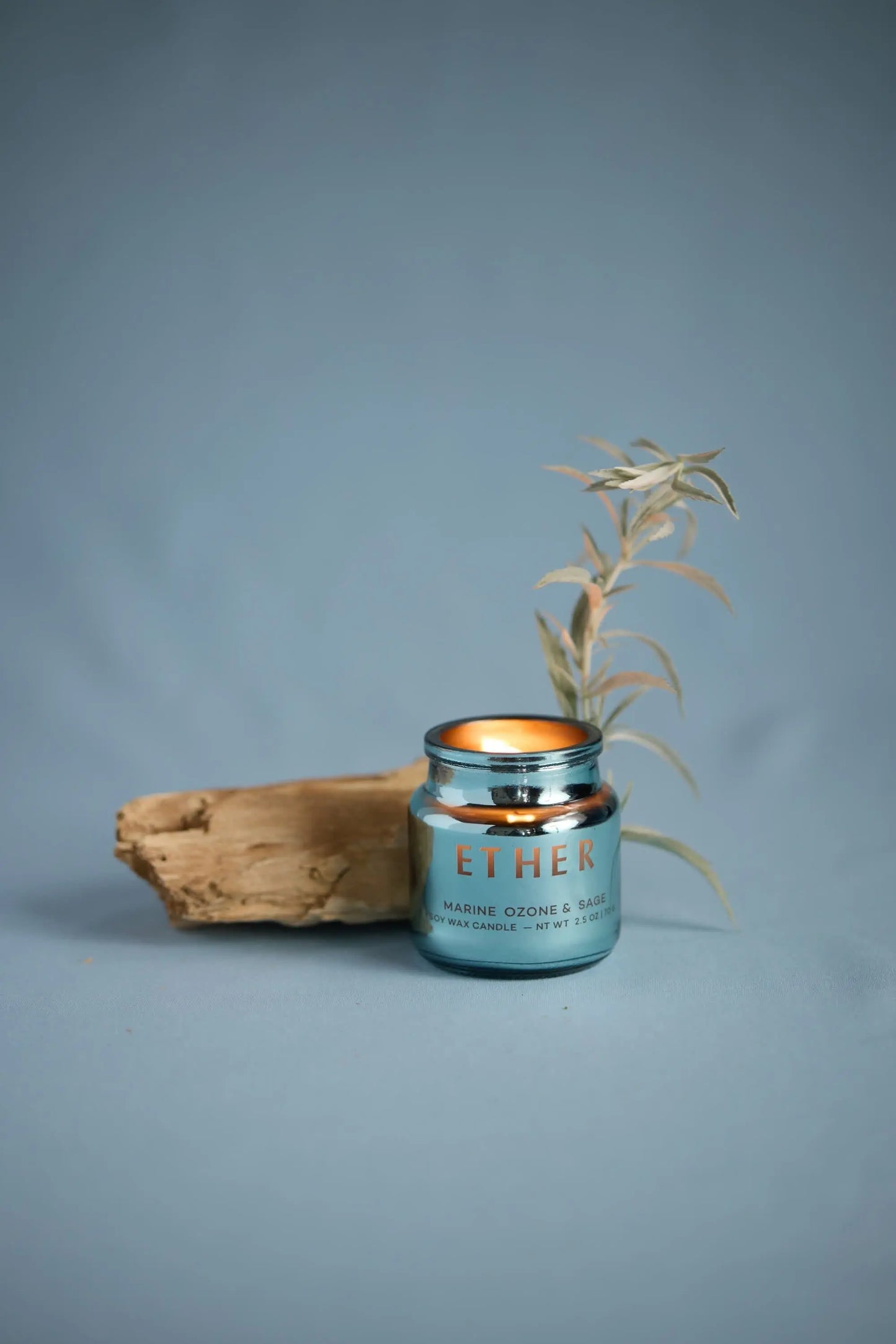 A metallic blue Chroma Candle - Sea Salt & Sage sits in its electroplated glass beside driftwood and dried foliage on a blue background.