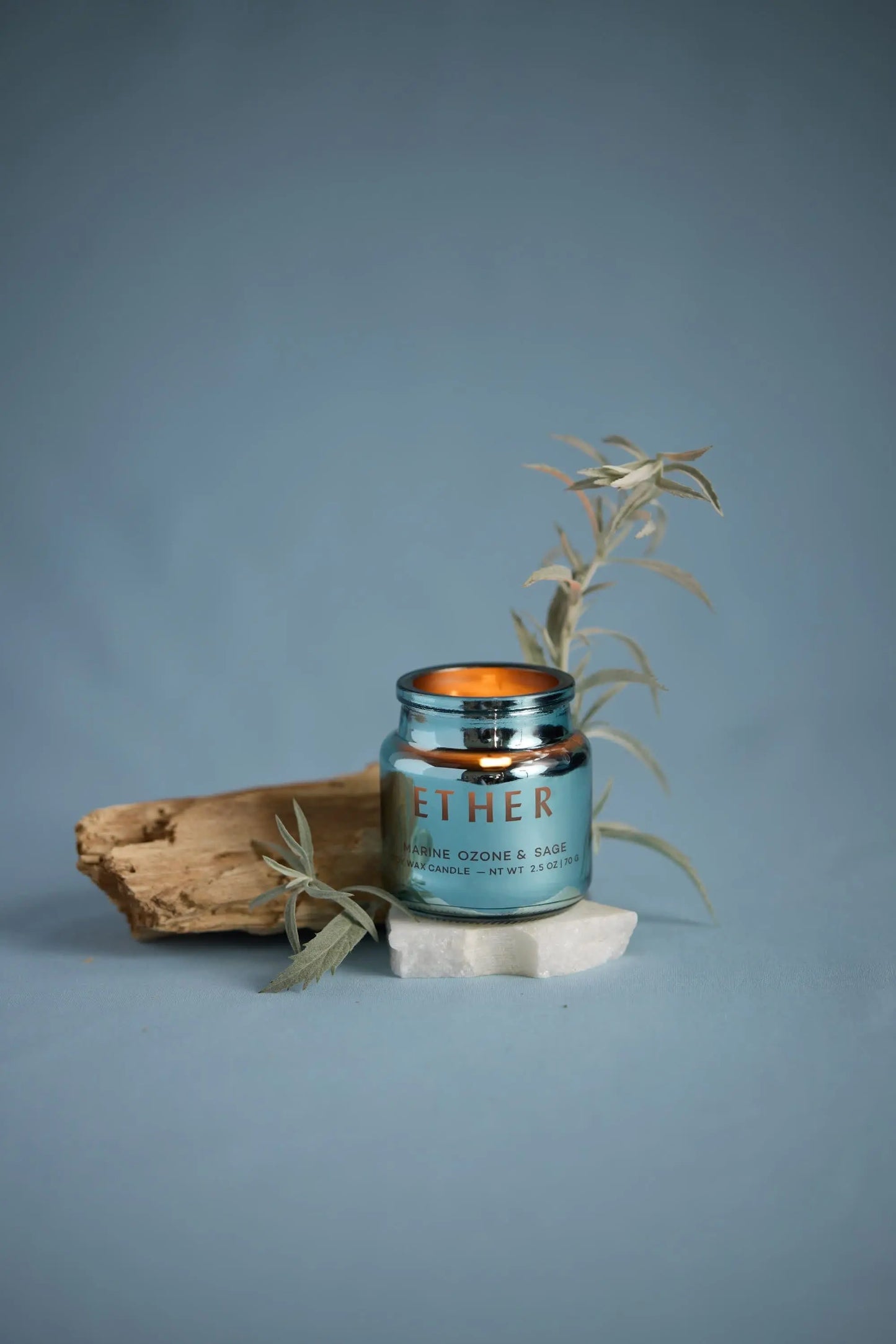 A blue electroplated glass Chroma Candle - Sea Salt & Sage sits on white stone with wood and foliage, capturing the refined style of the collection against a muted blue background.