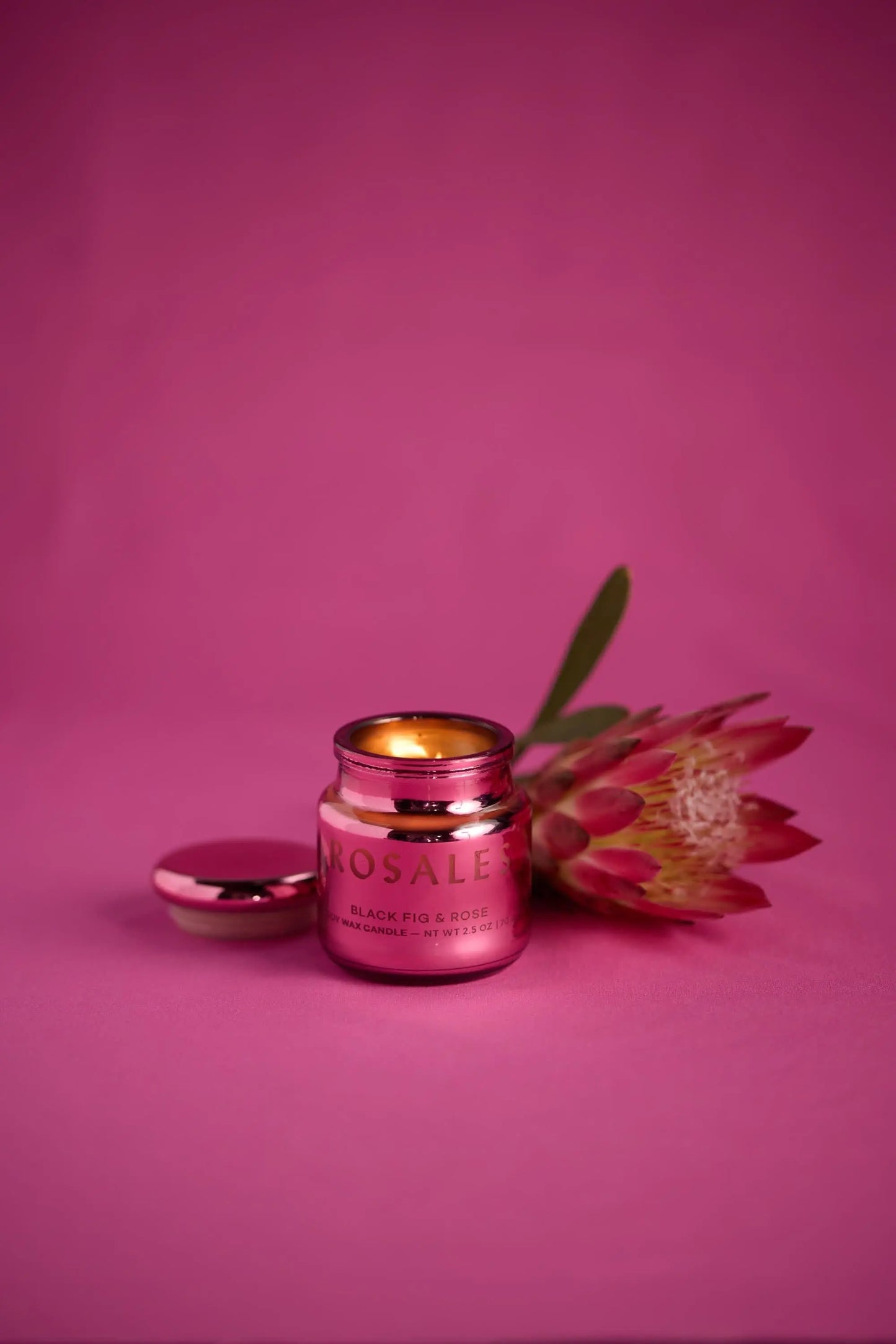 A small pink jar candle labeled CHROMA CANDLE - SAFFRON ROSE sits open with a lit flame next to a single protea flower, against a pink background—part of the Chroma collection for a unique scent experience.