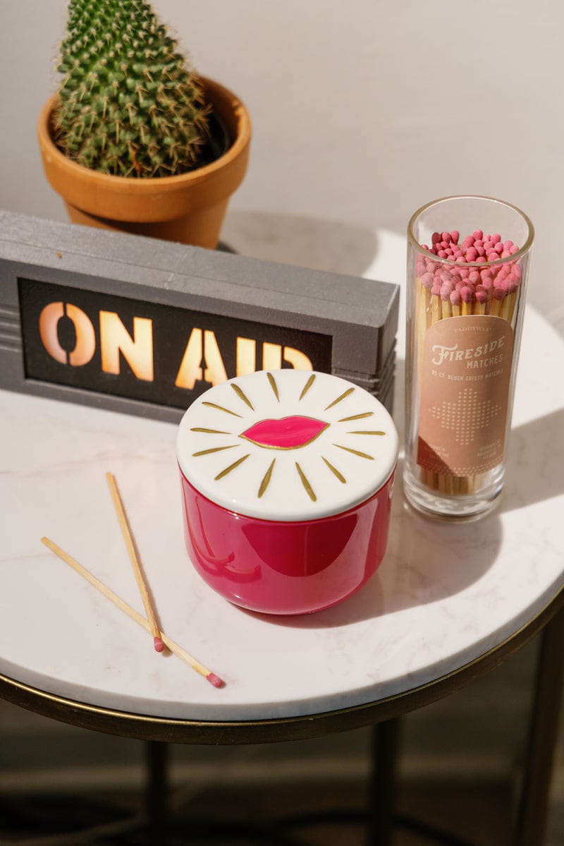 A small round table displays a potted cactus, an ON AIR sign, a lips-decorated red jar with lid, pink-tipped matches in glass, and the Smoked Cherry - Charmed 8oz Candle—an ideal gift for any occasion.