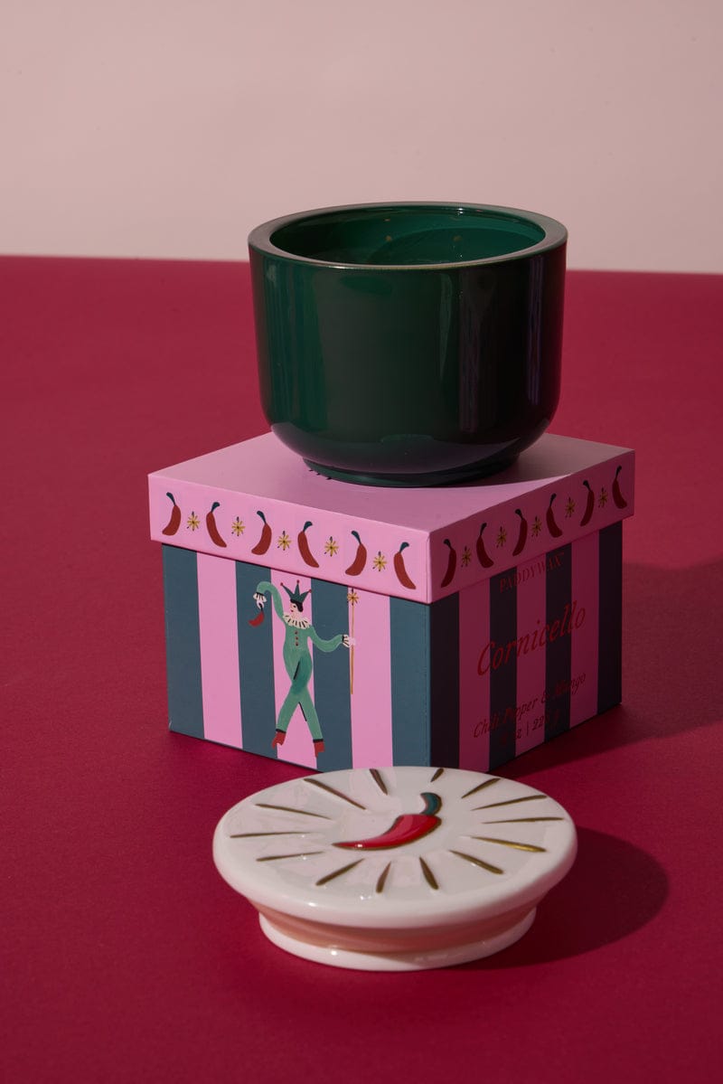The Chili Pepper & Mango - Charmed 8oz Candle sits on a pink striped box featuring a circus performer; its white ceramic lid, decorated with a red chili pepper, is displayed in the foreground.