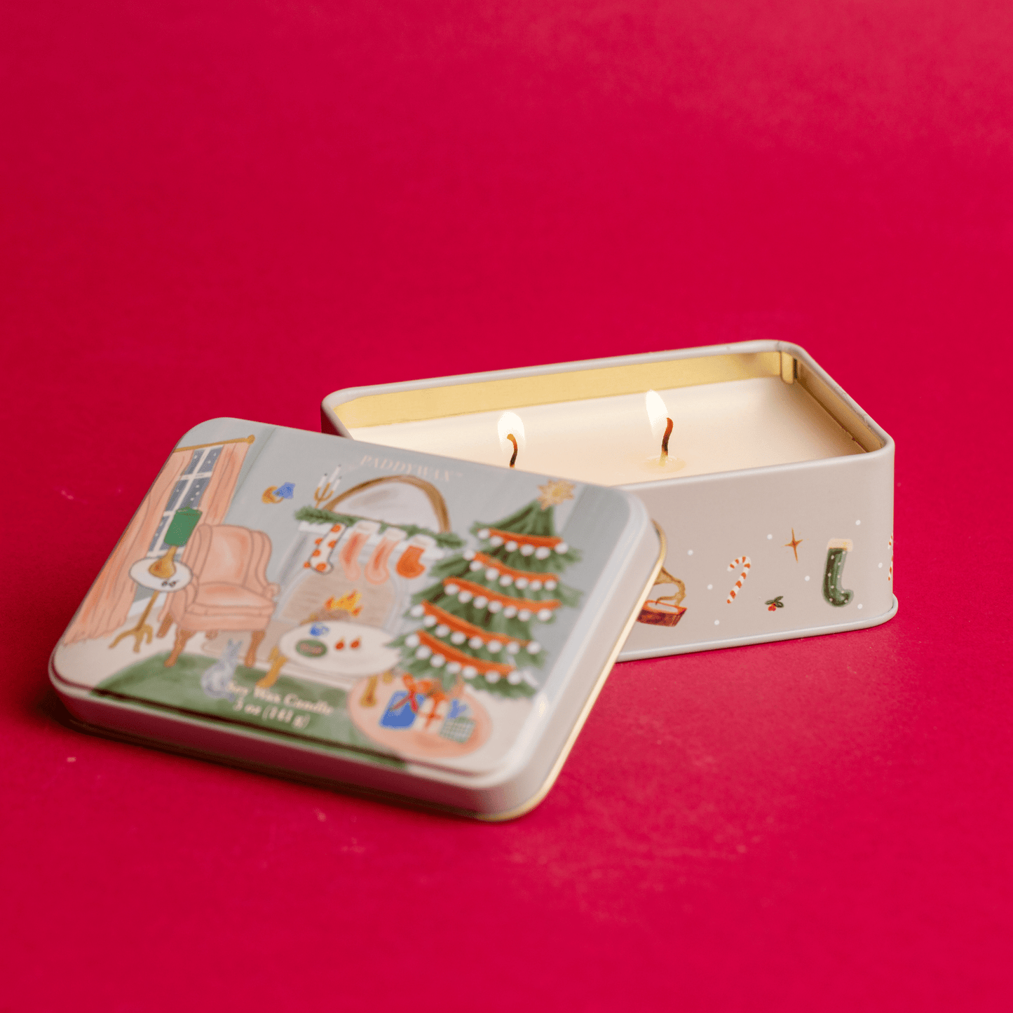 Front view of 5 oz holiday tin with two wicks and custom artwork resting on red table and lit
