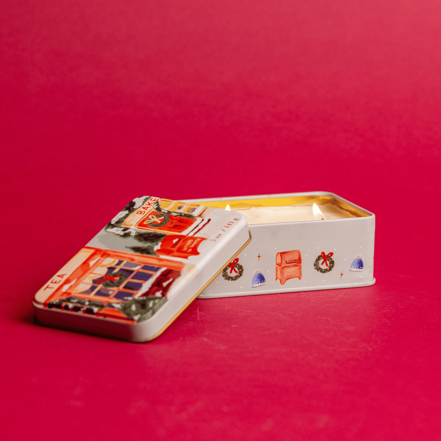 Front view of 5 oz holiday tin with custom artwork; lid shows snow-covered storefronts; on red table
