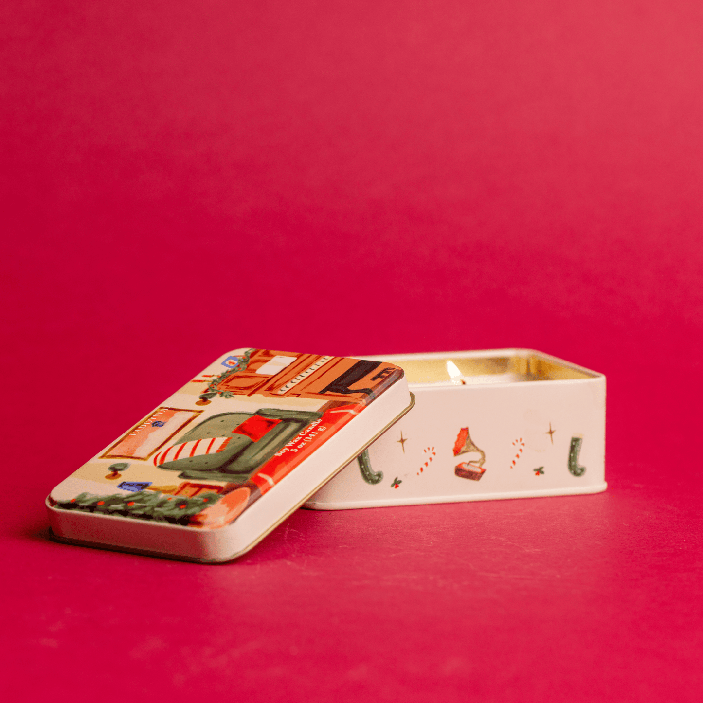 On a red background, a lit two wick 5 oz holiday tin with custom artwork; lid shows family room with piano and decorated tree