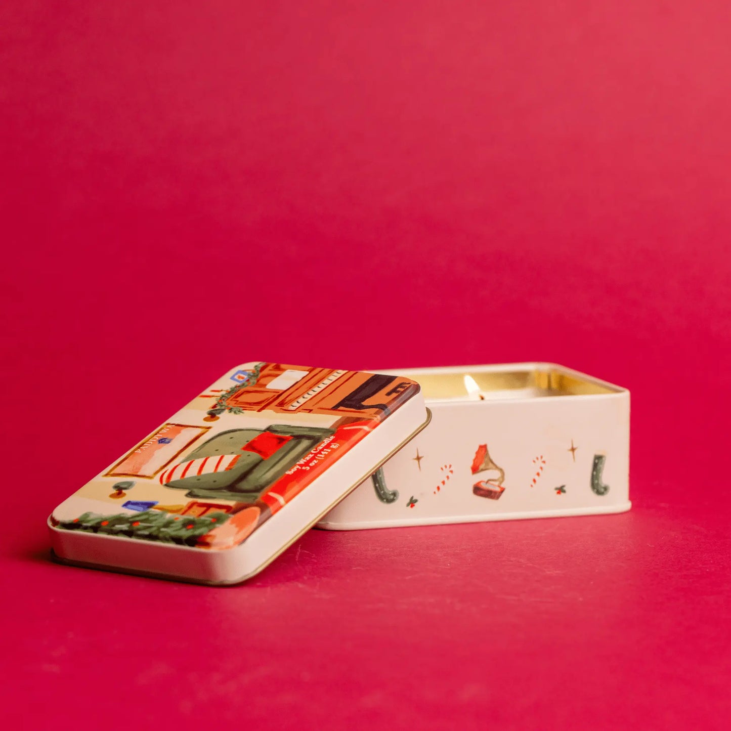 On a red background, a lit two wick 5 oz holiday tin with custom artwork; lid shows family room with piano and decorated tree