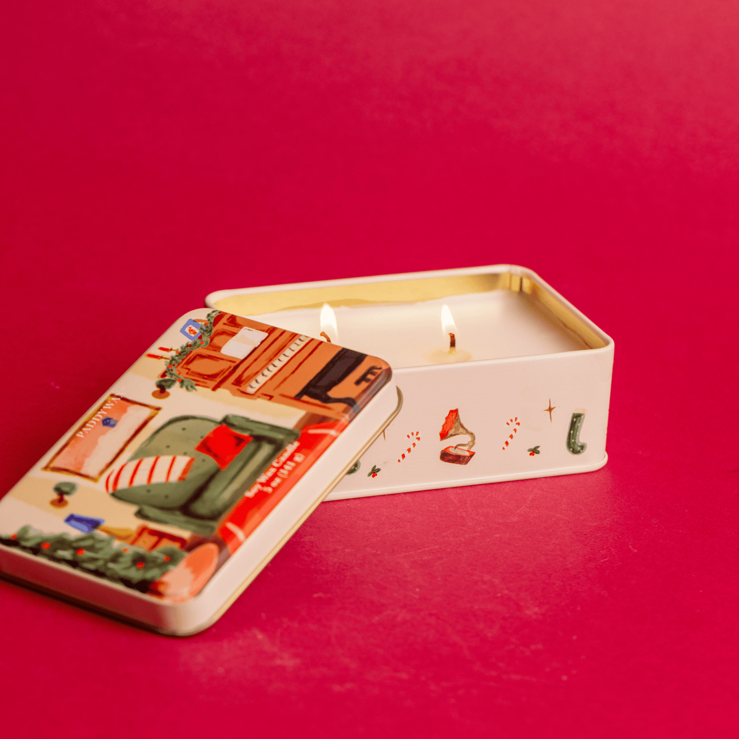On a red background, a top view of a two wick, lit 5 oz holiday tin with custom artwork; lid shows family room with piano and decorated tree