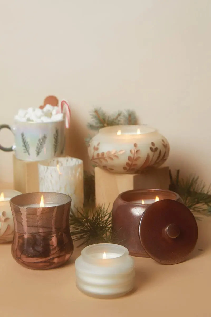 A Cypress & Fir 9oz frosted white glass candle with copper metallic branch etching sits on a beige surface beside pine branches and a mug of hot chocolate topped with marshmallows and a candy cane, creating cozy holiday decor.