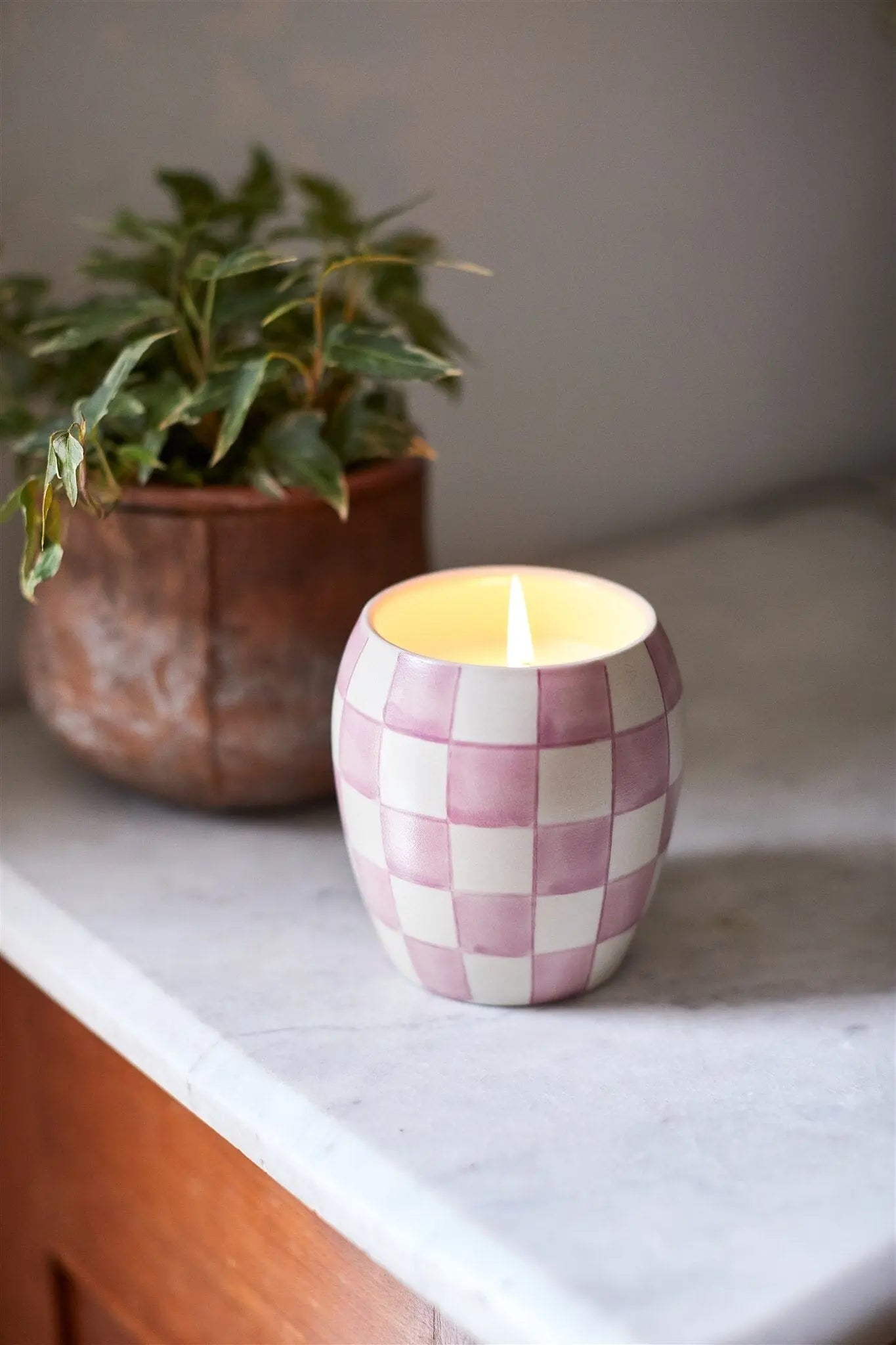 The Checkmate 11 oz Candle - Lavender Mimosa in a pink and white checkerboard holder sits on marble beside a green plant, adding cozy light and reusable decor.