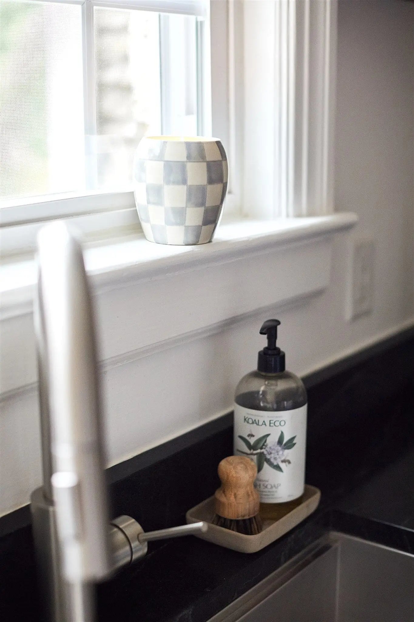 A Checkmate 11 oz Candle - Cotton + Teak with a classic checkerboard print sits on a windowsill above a kitchen sink, alongside Koala Eco dish soap and two scrub brushes in a tray on the counter.