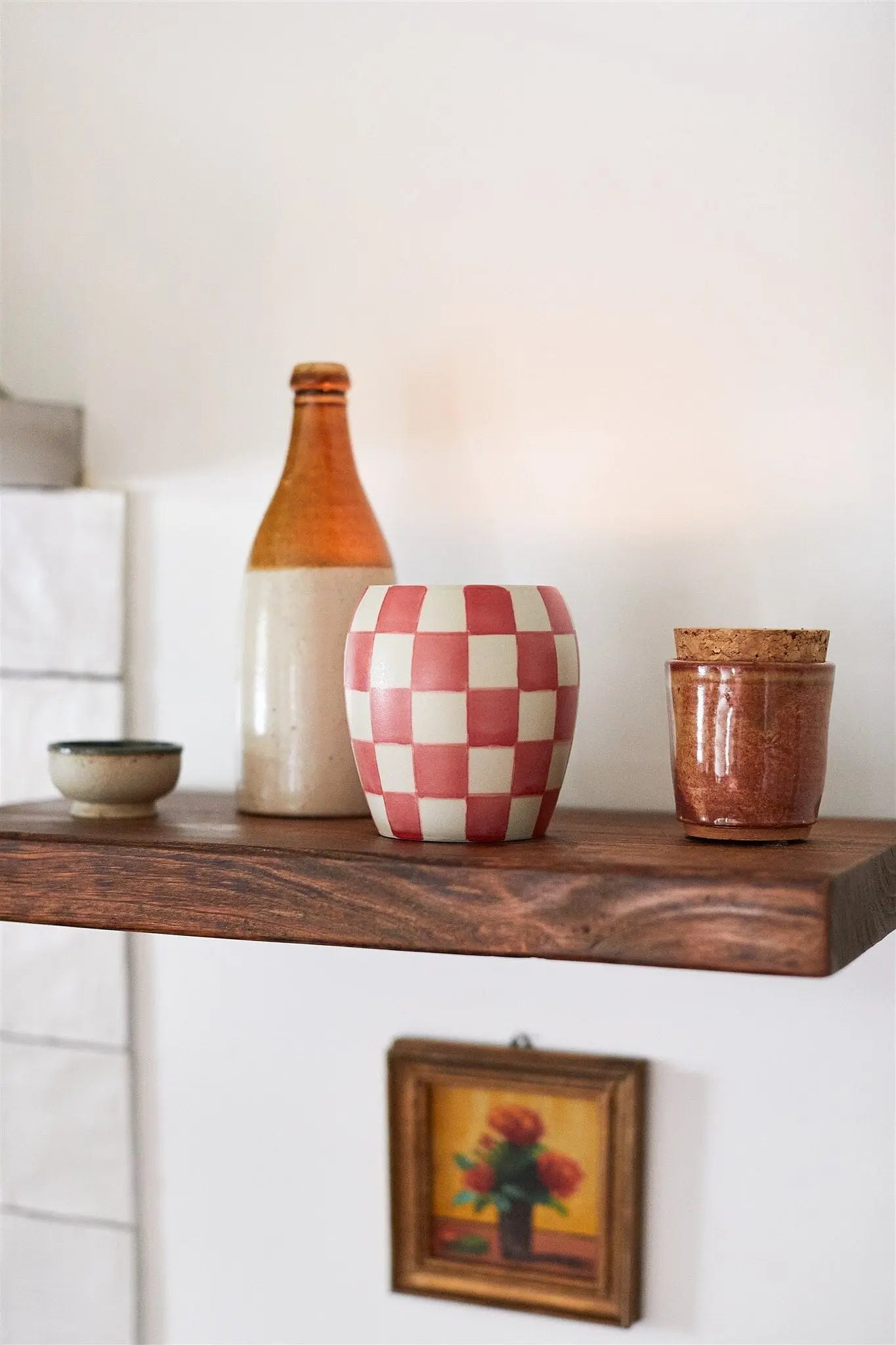 A wooden shelf displays hand-painted vessels, including the Checkmate 11 oz Candle - Rose + Santal, a brown bottle, a small bowl, a jar with a cork lid, and a framed picture on the wall below.
