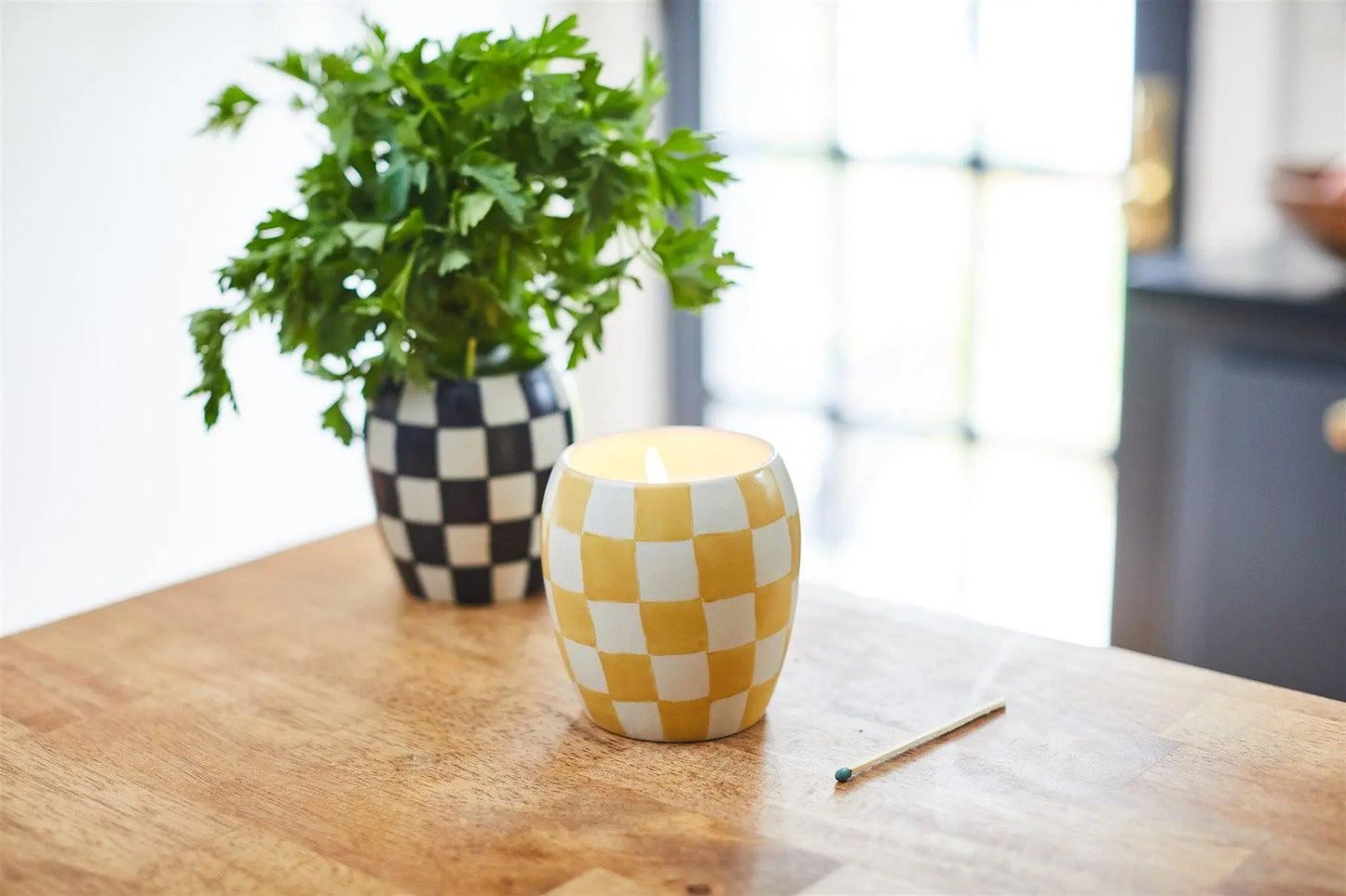 The Checkmate 11 oz Candle - Golden Amber, in a yellow and white checkerboard holder, sits on a wooden table beside a matchstick and a vase with green leaves in a hand-painted vessel.