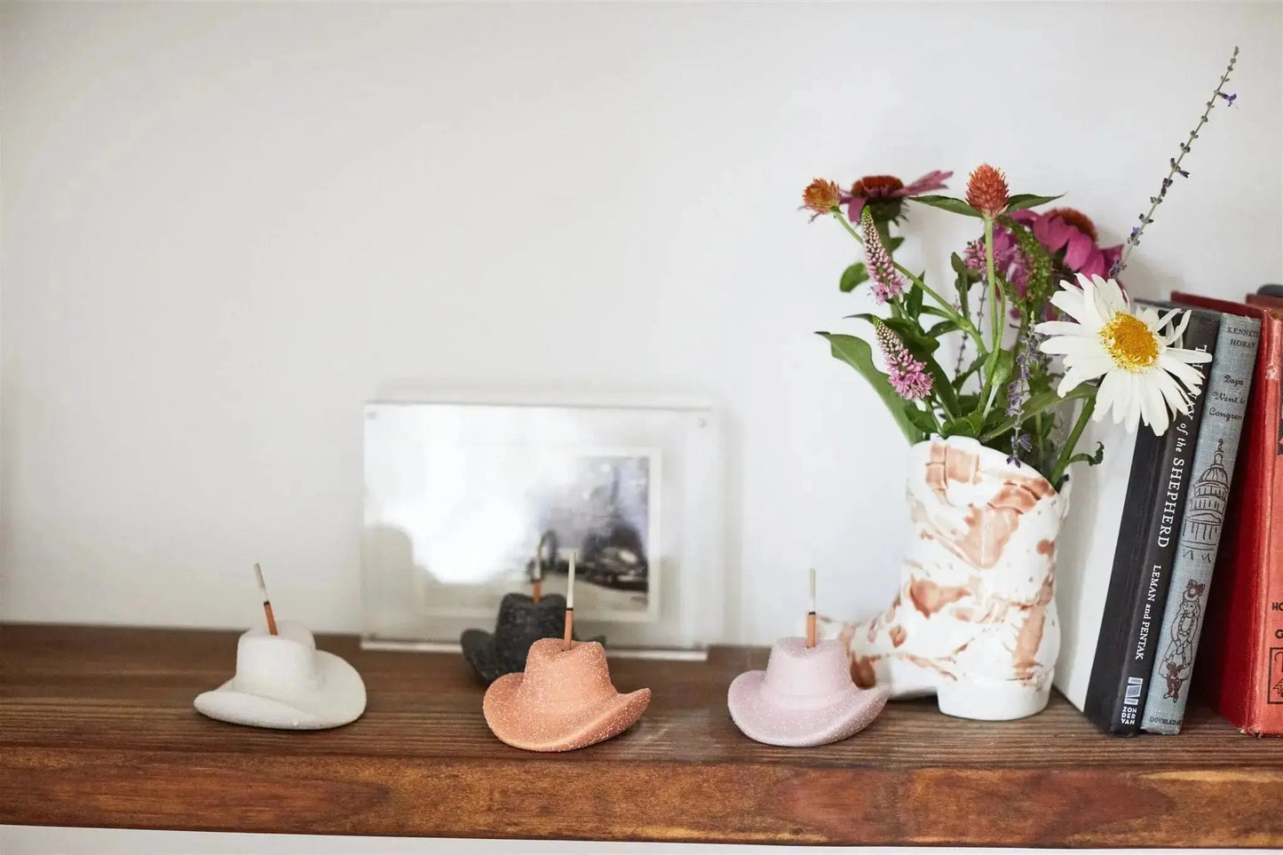Four Cowboy Hat Incense Holder - Black are displayed on a wooden shelf beside a marbled vase with flowers, framed art, and a row of books.