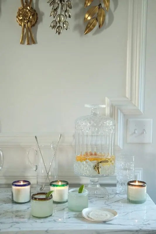 La Playa 9 oz Candle - Salted Blue Agave sits next to a hand-blown bubble glass beverage dispenser, two drink glasses, and recycled glassware on a marble countertop; gold wall decor above. Inspired by La Playa Recipe Blog.