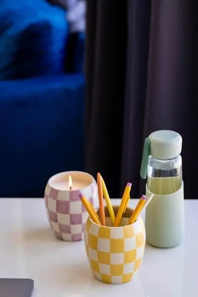 A desk with a Checkmate 11 oz Candle - Golden Amber lit, a yellow and white checkerboard cup holding pencils, a light green water bottle, and a laptop.