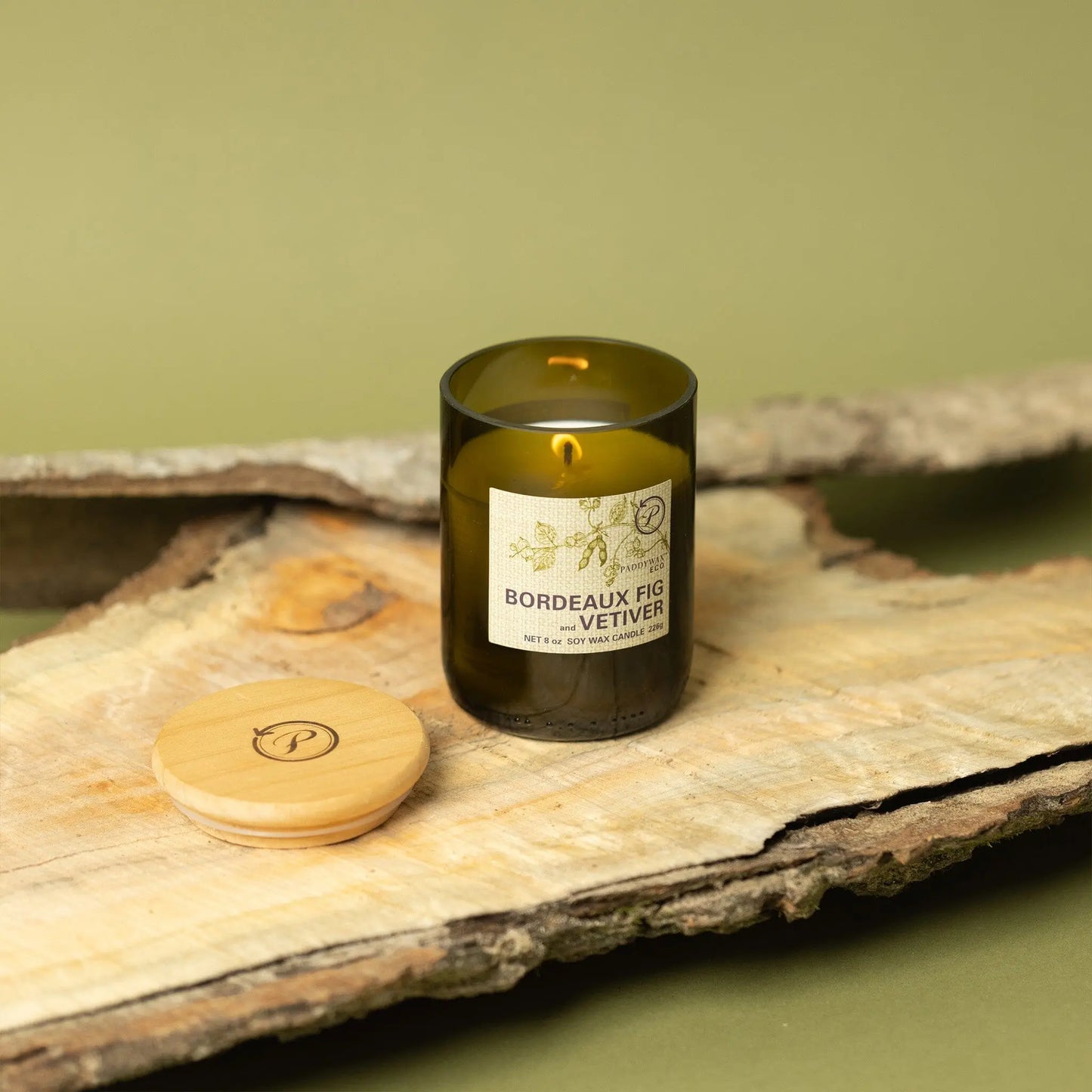 The Eco 8 oz Candle - Bordeaux Fig + Vetiver in a green glass jar rests on wood with its wooden lid nearby, set against a neutral green background—ideal for anyone looking for sustainable home decor.