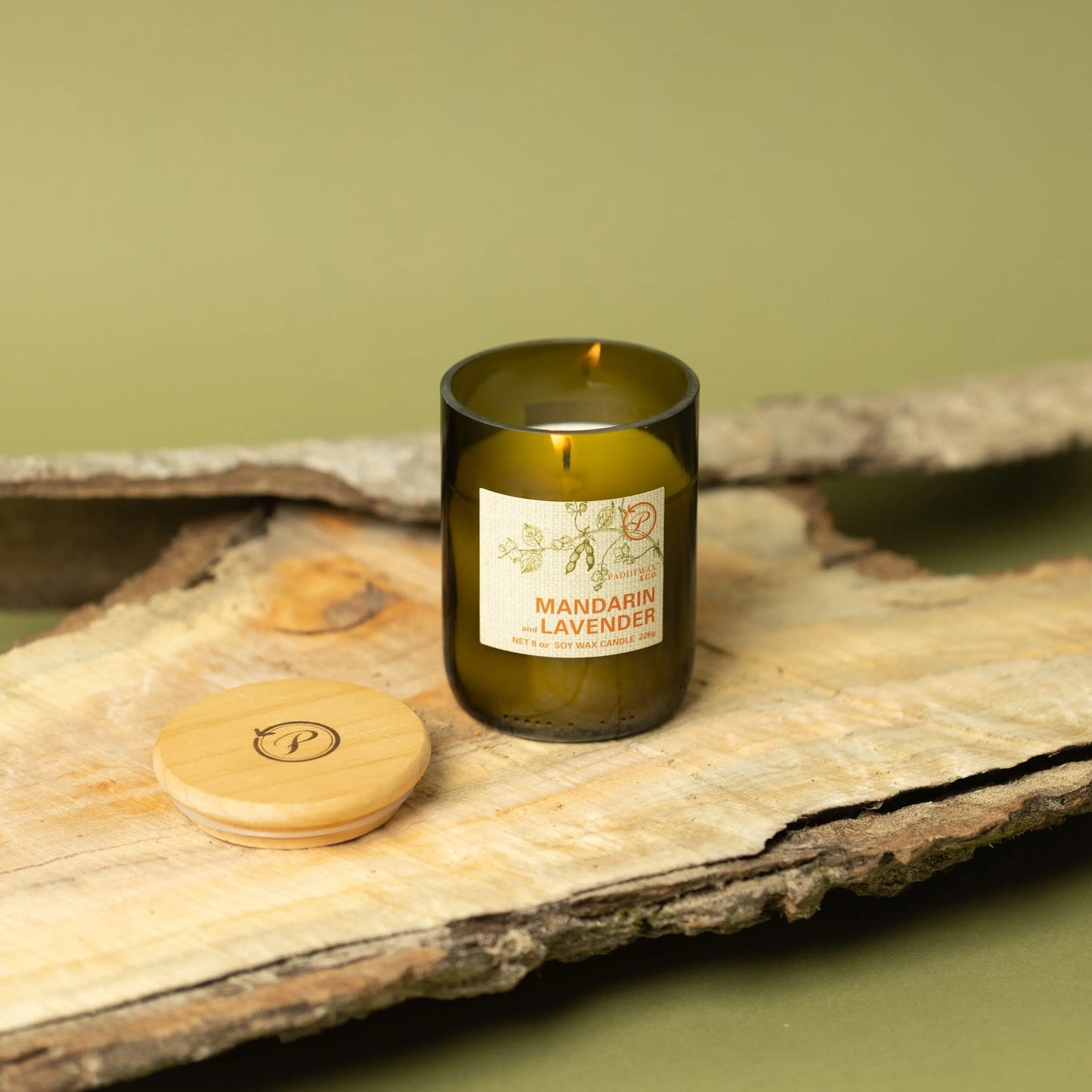 The Mandarin + Lavender - Eco 8 oz Candle, in a dark green glass jar with a round wooden lid, sits on wood against a green background—an inviting accent for sustainable living.