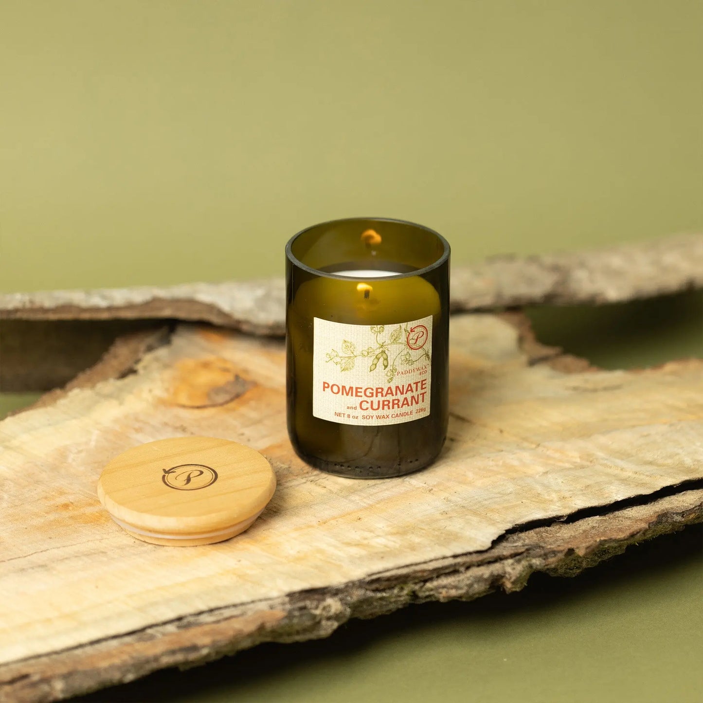 The Pomegranate + Currant - Eco 8 oz Candle, a green glass candle with two wicks and a botanical label, rests on wood with its wooden lid nearby, set against an olive green background.
