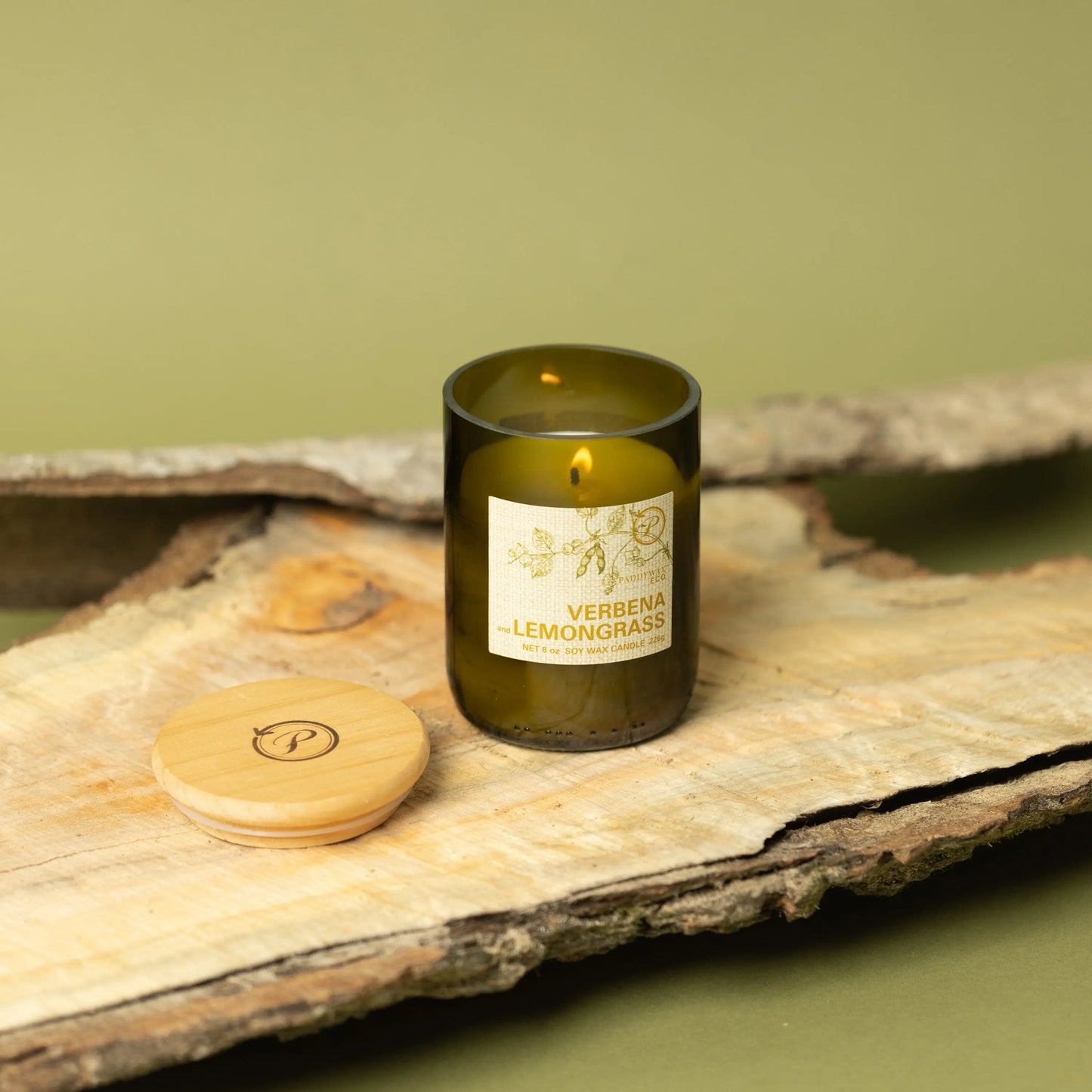 The Eco 8 oz Candle – Verbena + Lemongrass, in a green glass jar with a wooden lid, sits on wood against a green backdrop—ideal for repurposing or adding sustainable style to your space.