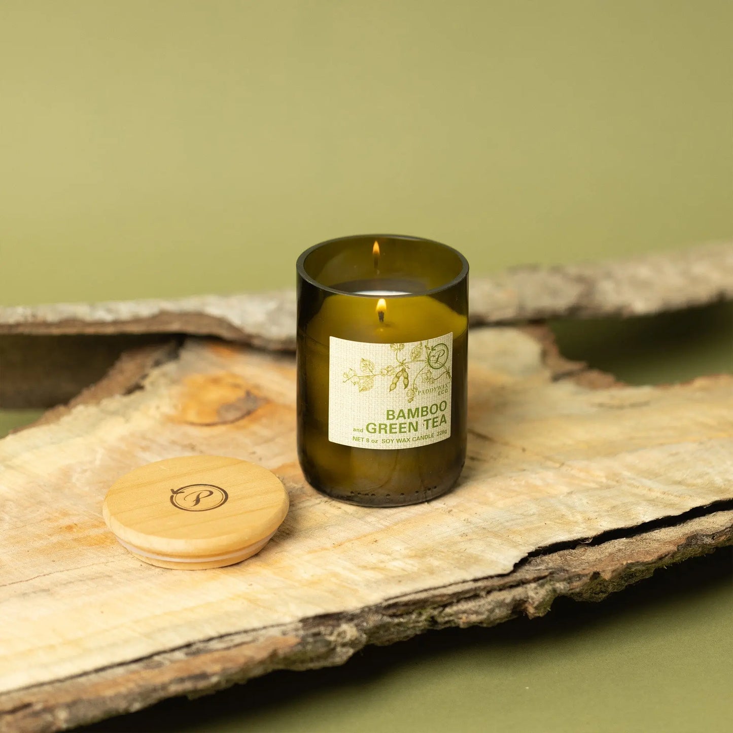 The Bamboo + Green Tea - Eco 8 oz Candle, in a glass jar with a wooden lid, sits on wood against a green background—an inviting accent for anyone who values sustainable living.