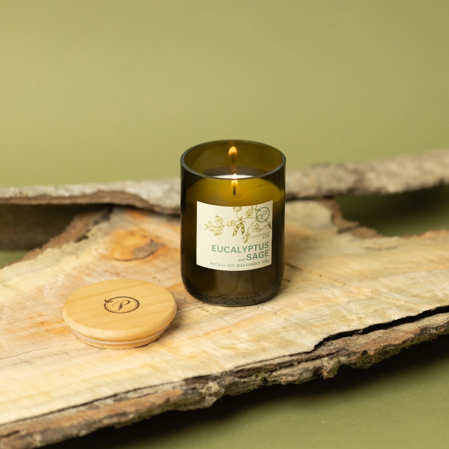 The Eco 8 oz Candle - Eucalyptus + Sage glows from a dark recycled vessel on wood, its wooden lid beside it, set against green—a stylishly sustainable choice for your home.