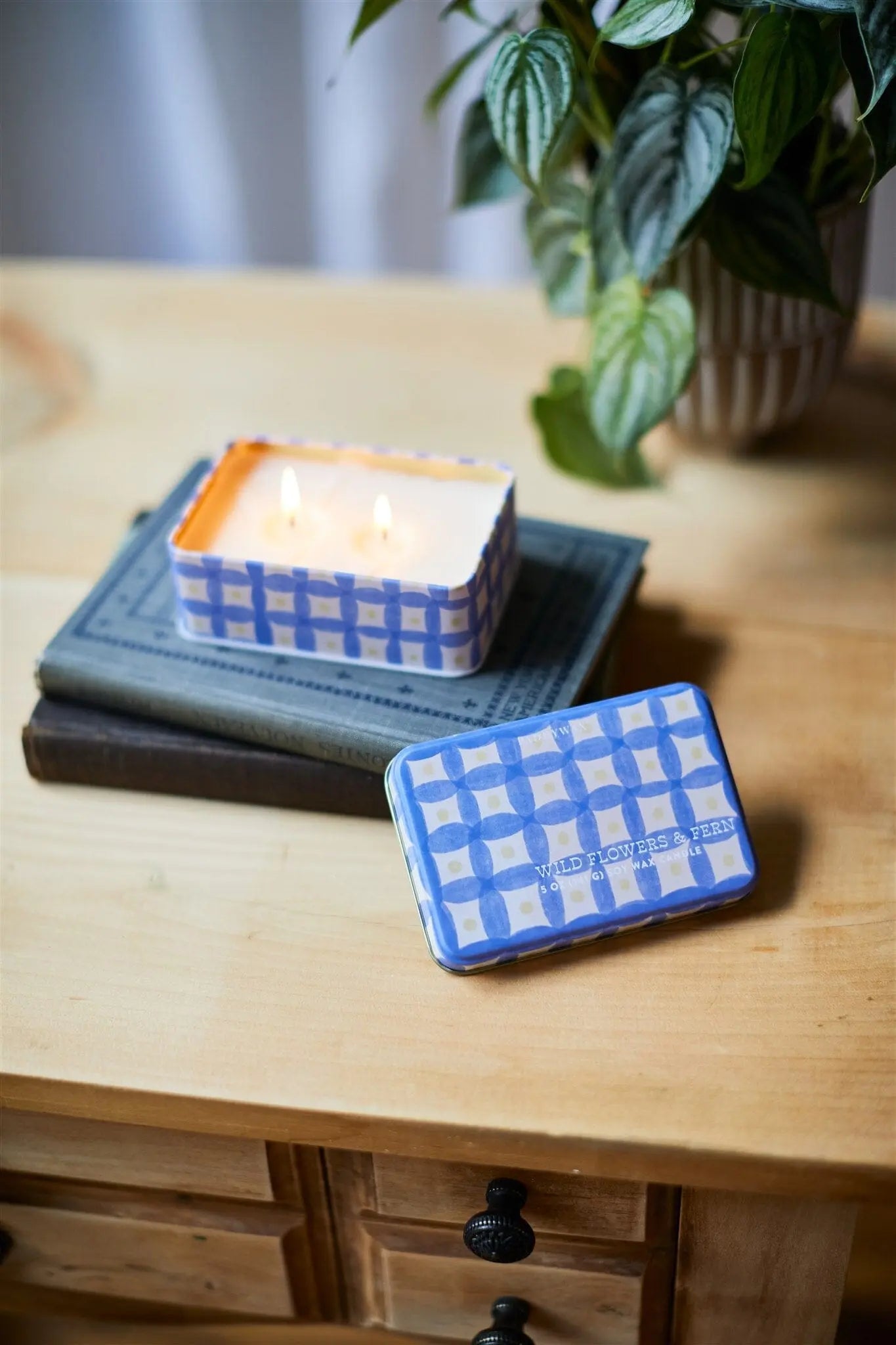 A lit Everyday Tins 5 oz. Candle - Wild Flowers & Fern, in a blue and white checkered tin, fills the air with fragrance as it sits on stacked books beside a plant on a wooden table, with the lid resting nearby.