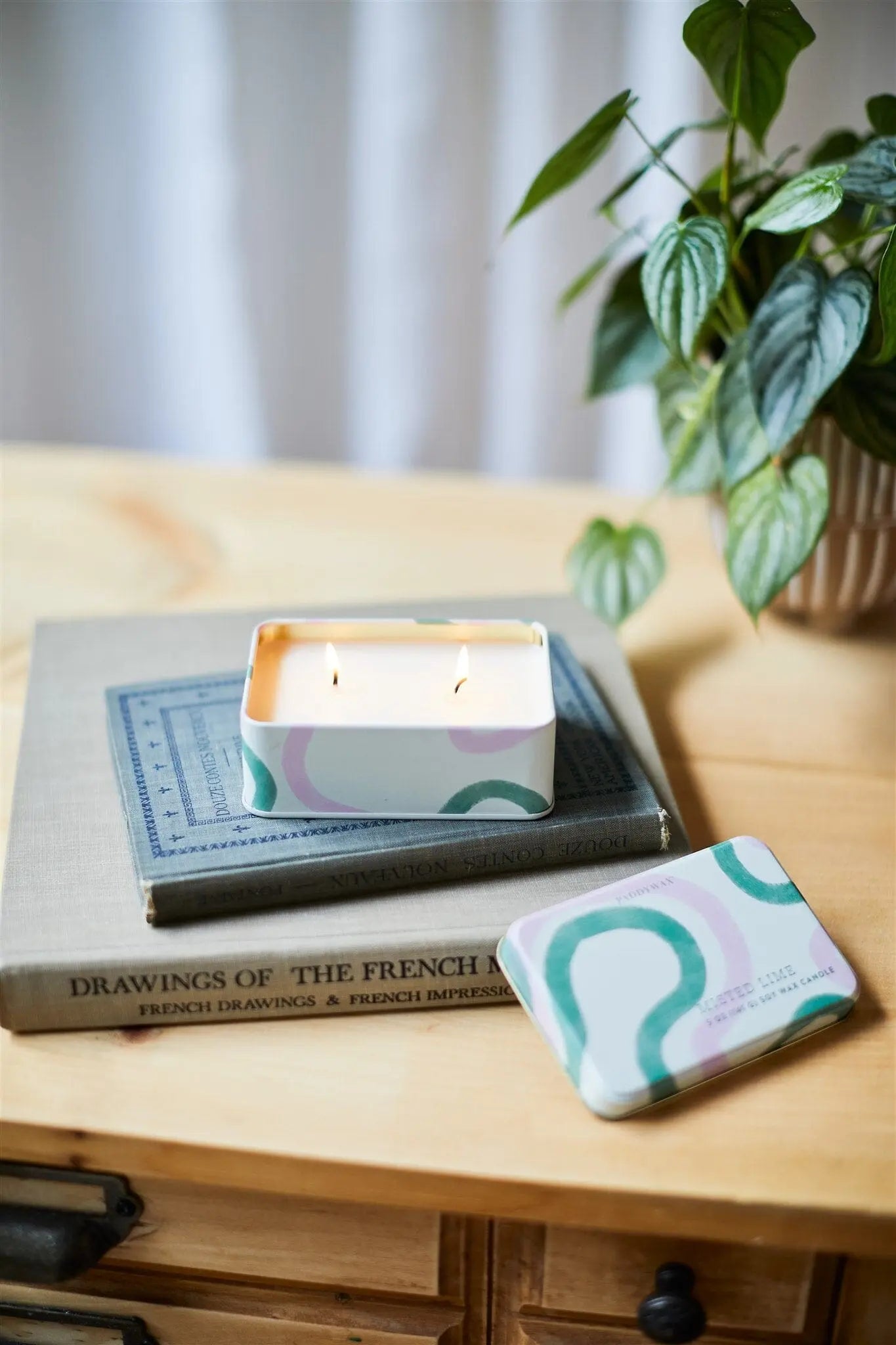 A lit Everyday Tins 5 oz. Candle - Misted Lime sits on stacked books beside its closed matching tin and a potted plant on a wooden table.